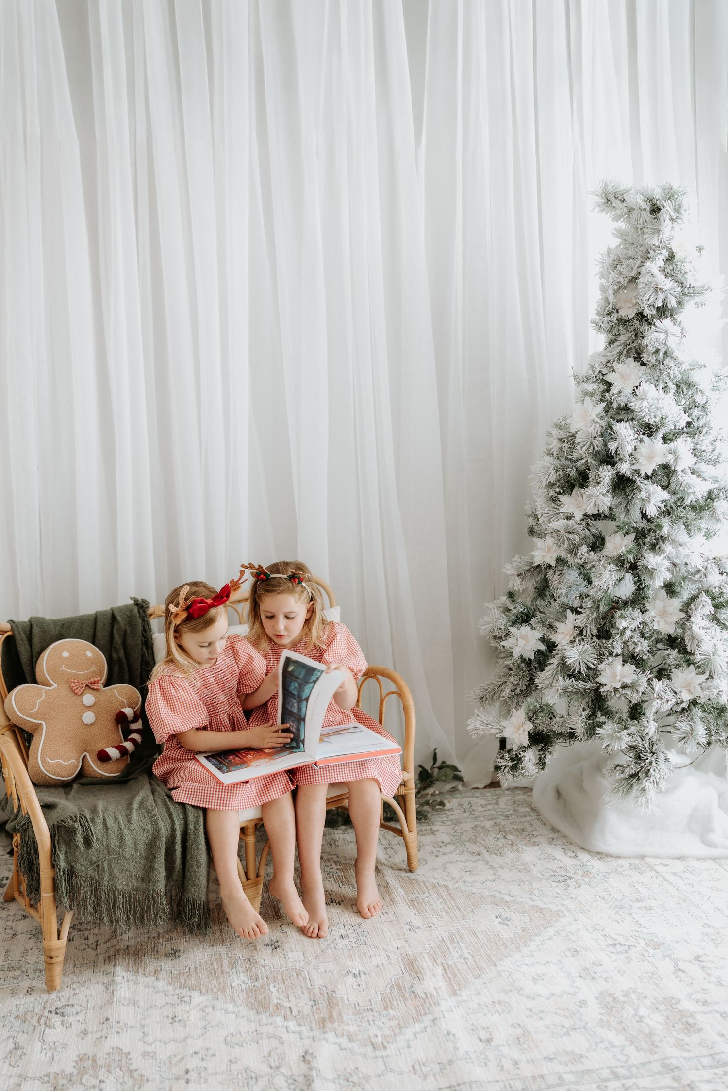 Two children in matching pink outfits sit on wooden chairs next to a flocked white Christmas tree in a studio setting.