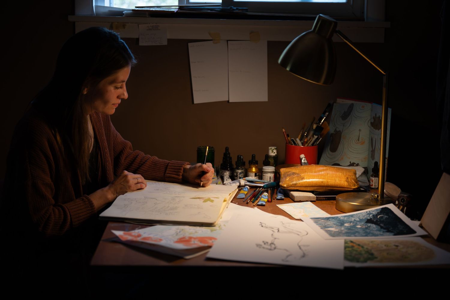 Artist working at illuminated desk in dark room sketches on paper with art supplies and desk lamp.