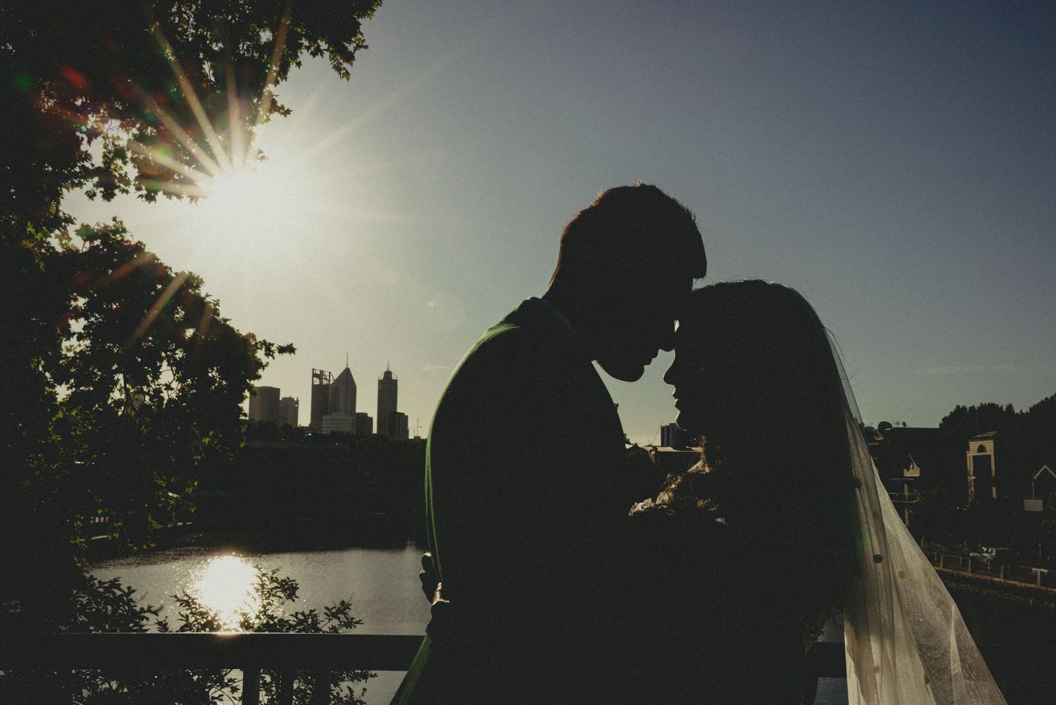 Silhouetted couple embraces during golden sunset by a lake with trees in the background.