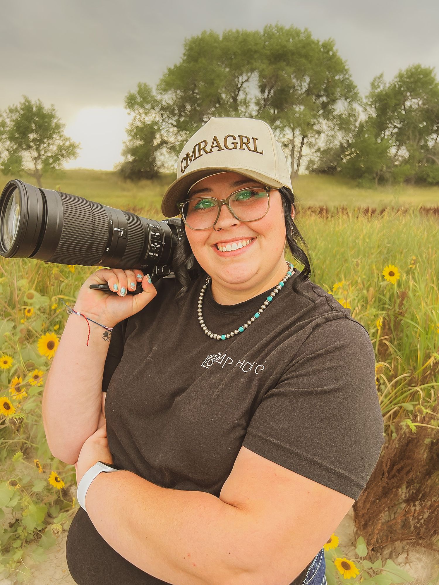 Photographer in sunflower field wearing a baseball cap and holding professional camera equipment.
