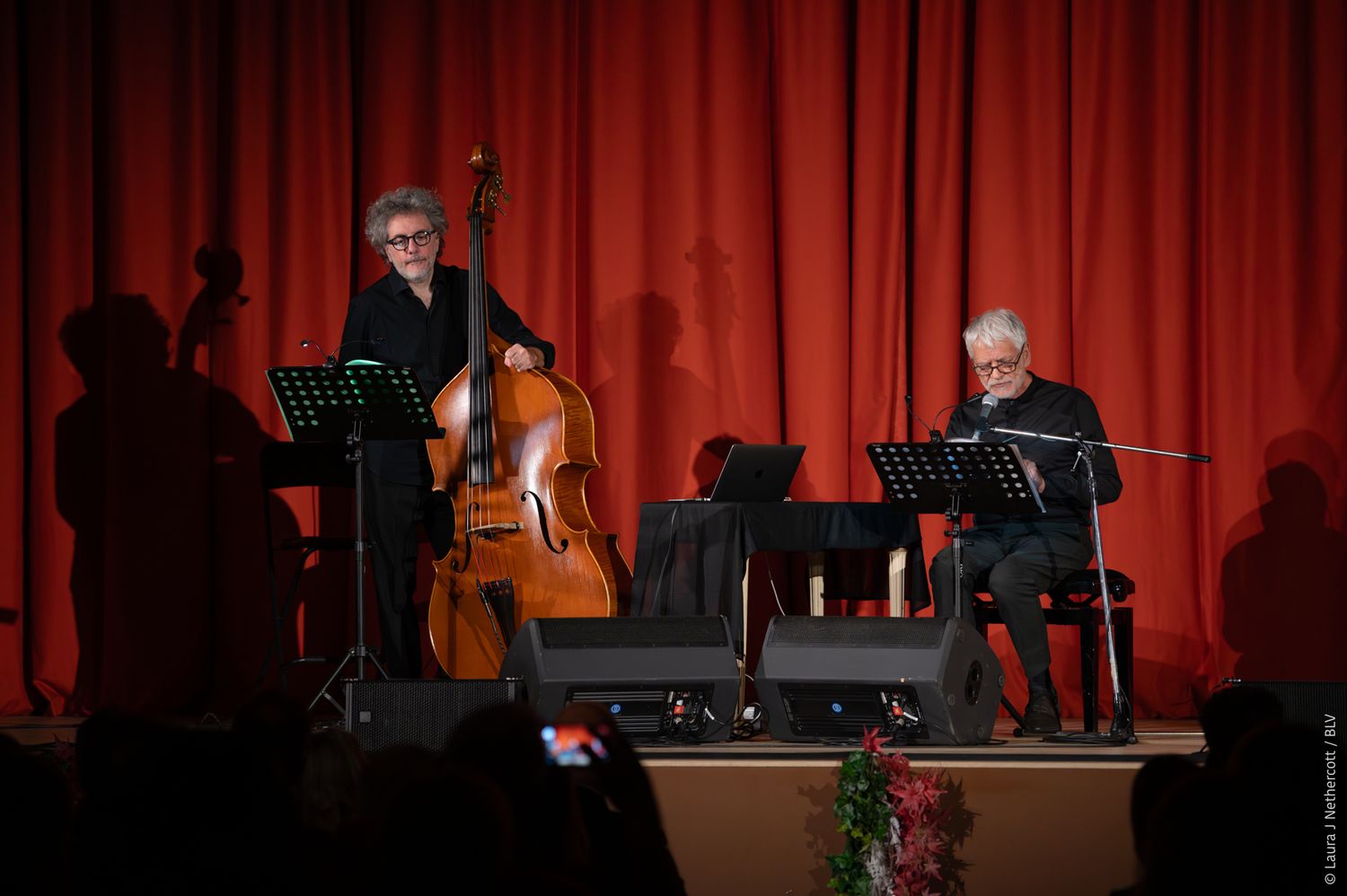 Musicians perform on stage with red curtain backdrop and string instruments visible in low light conditions.