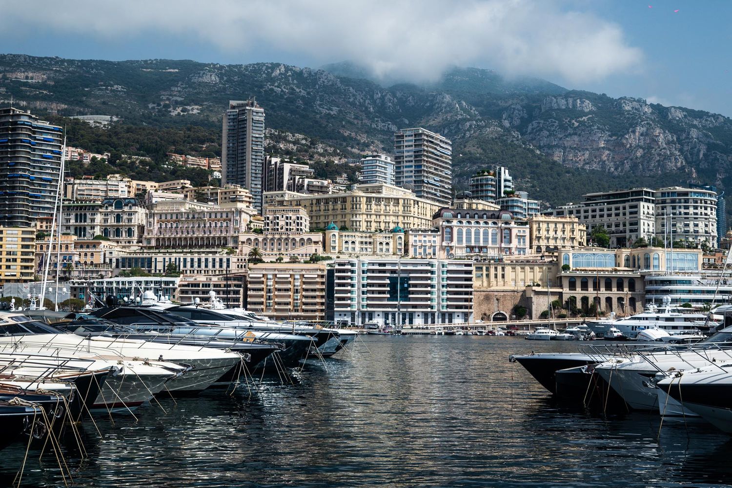 Luxury yachts docked in Monaco's harbor with high-rise buildings and mountains in the background.