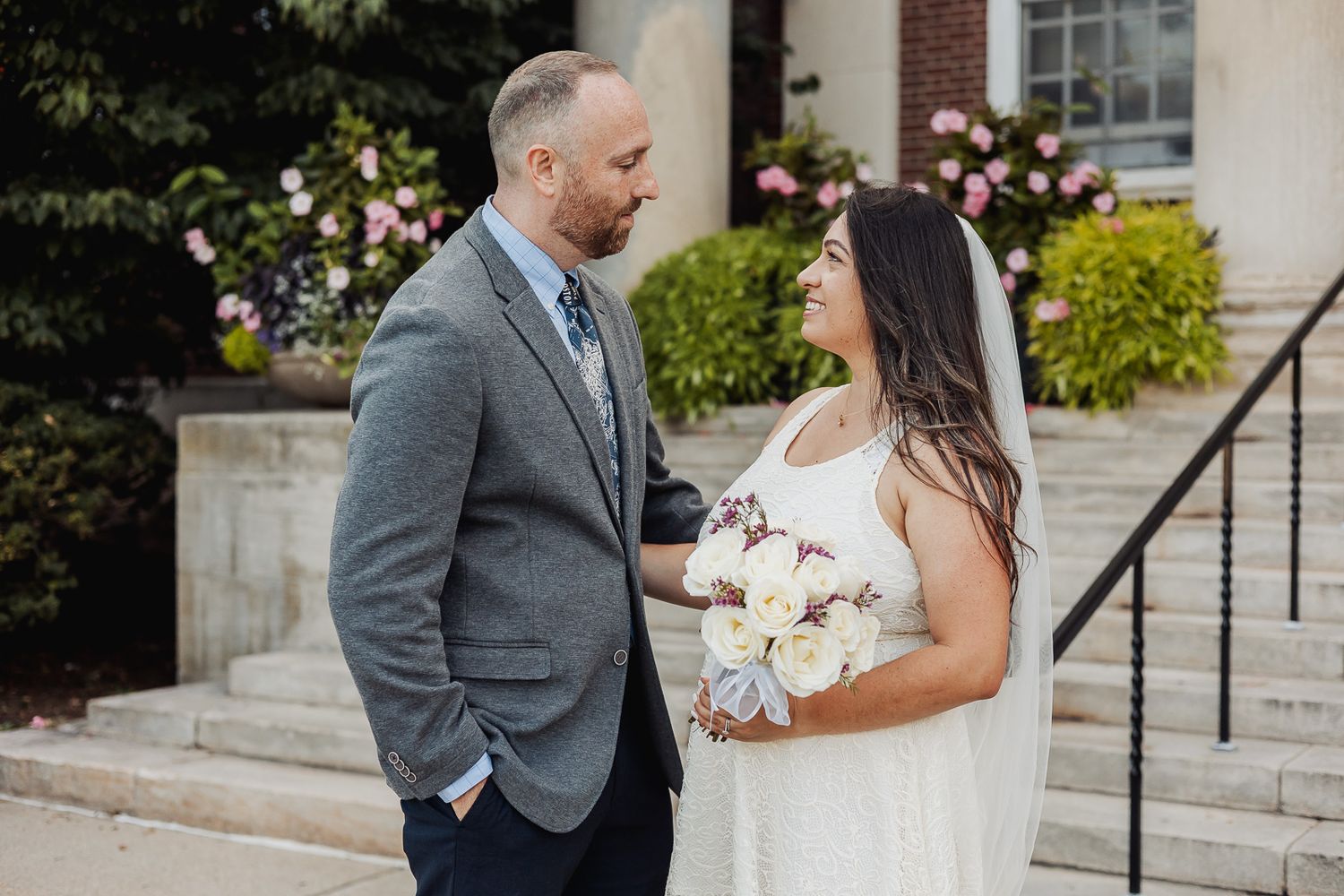 A couple shares romantic moments in their wedding attire on stone steps surrounded by blooming flowers.