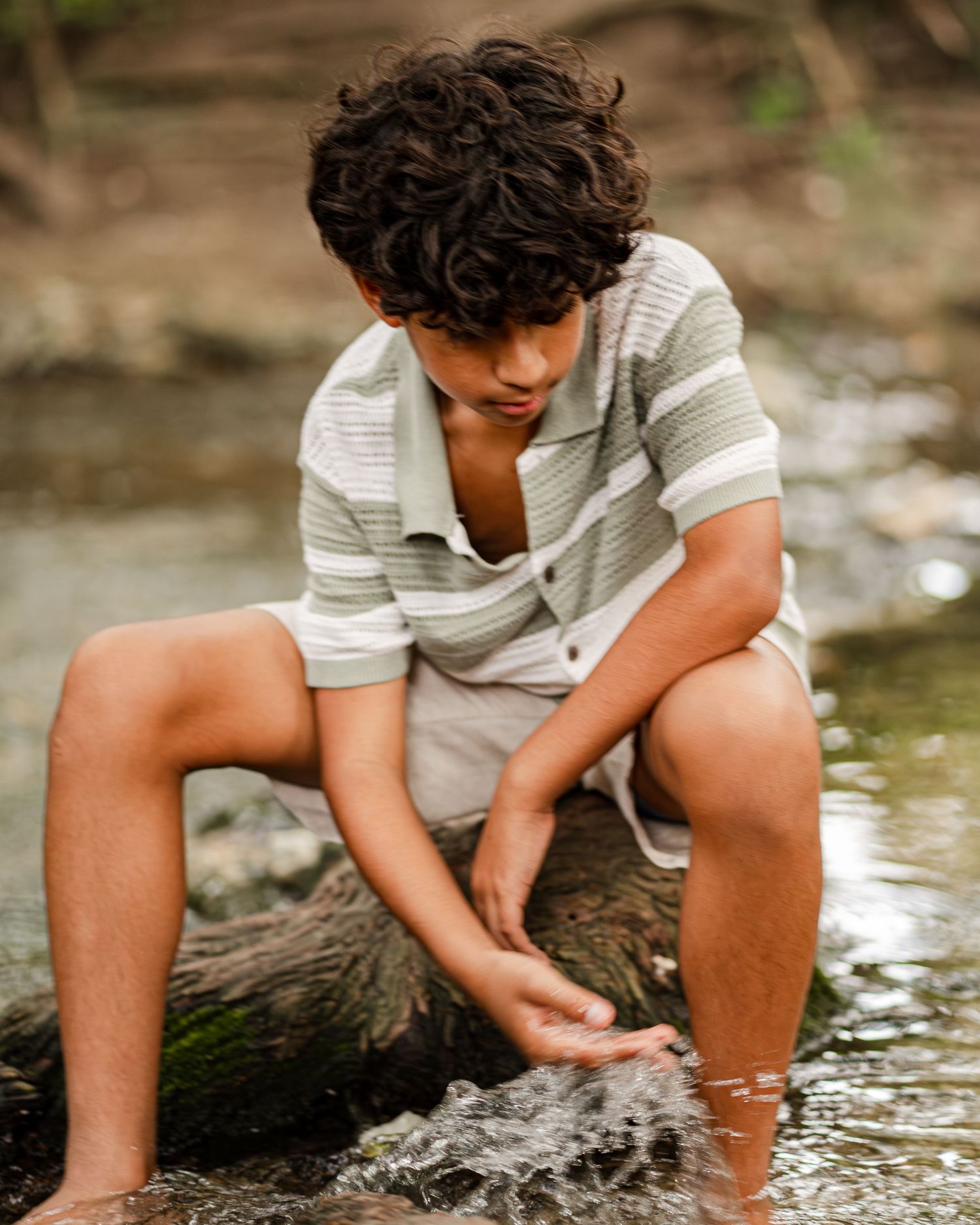 A person in a striped shirt sits on a rock by a stream, playing with the flowing water on a sunny day.