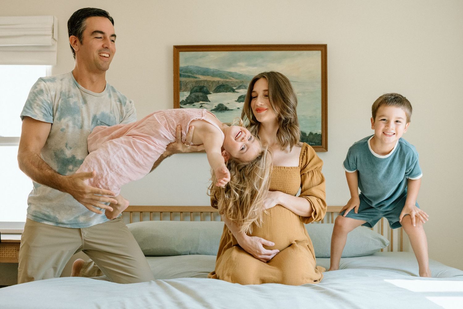 A cheerful family enjoys a playful moment together on the bed with a framed seascape painting on the wall.
