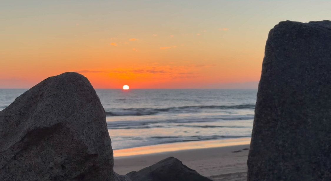 View of the sun setting over ocean with rocky outcroppings in foreground and dramatic orange sky at twilight.