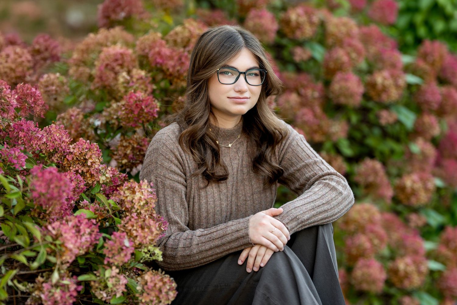 Teen girl with glasses sitting among pink hydrangea bushes in a brown knit sweater.