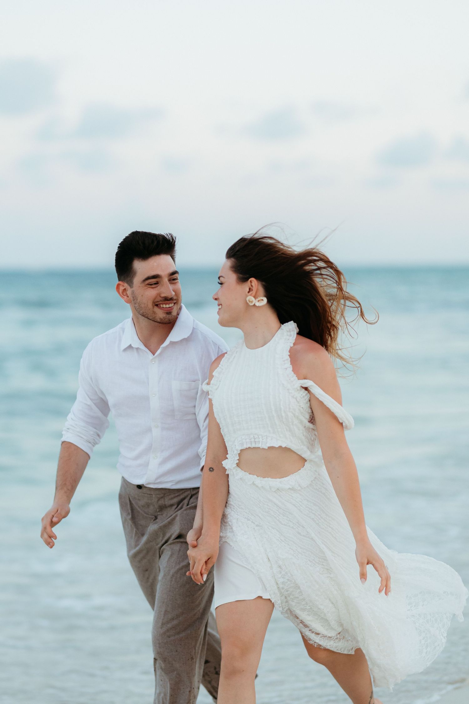 A couple in white and gray attire playfully runs along a beach with turquoise ocean waters in the background.
Tulum engagement photographer
