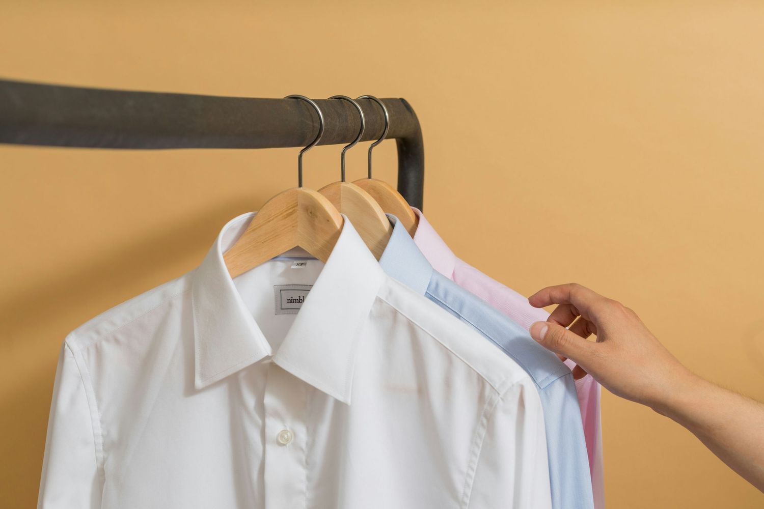 Hand reaching for white dress shirts hanging on wooden hangers against a beige wall.