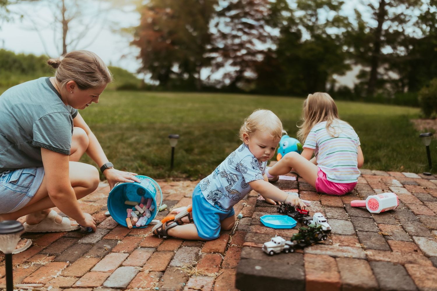 Children and adult play together with toys on a brick patio during a sunny afternoon outdoors.