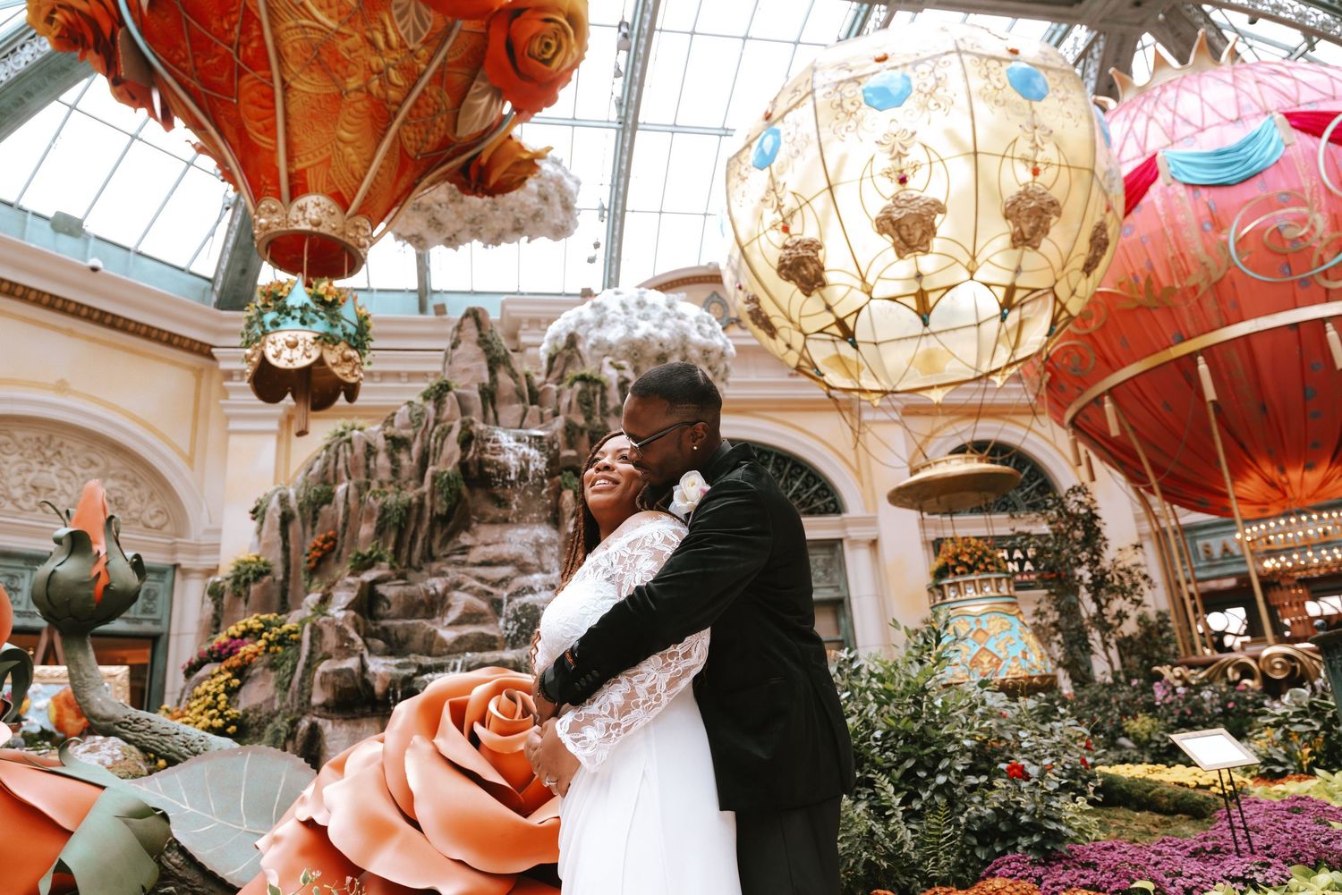 Two people embrace under ornate hanging lanterns and floral decorations in a luxurious conservatory setting.