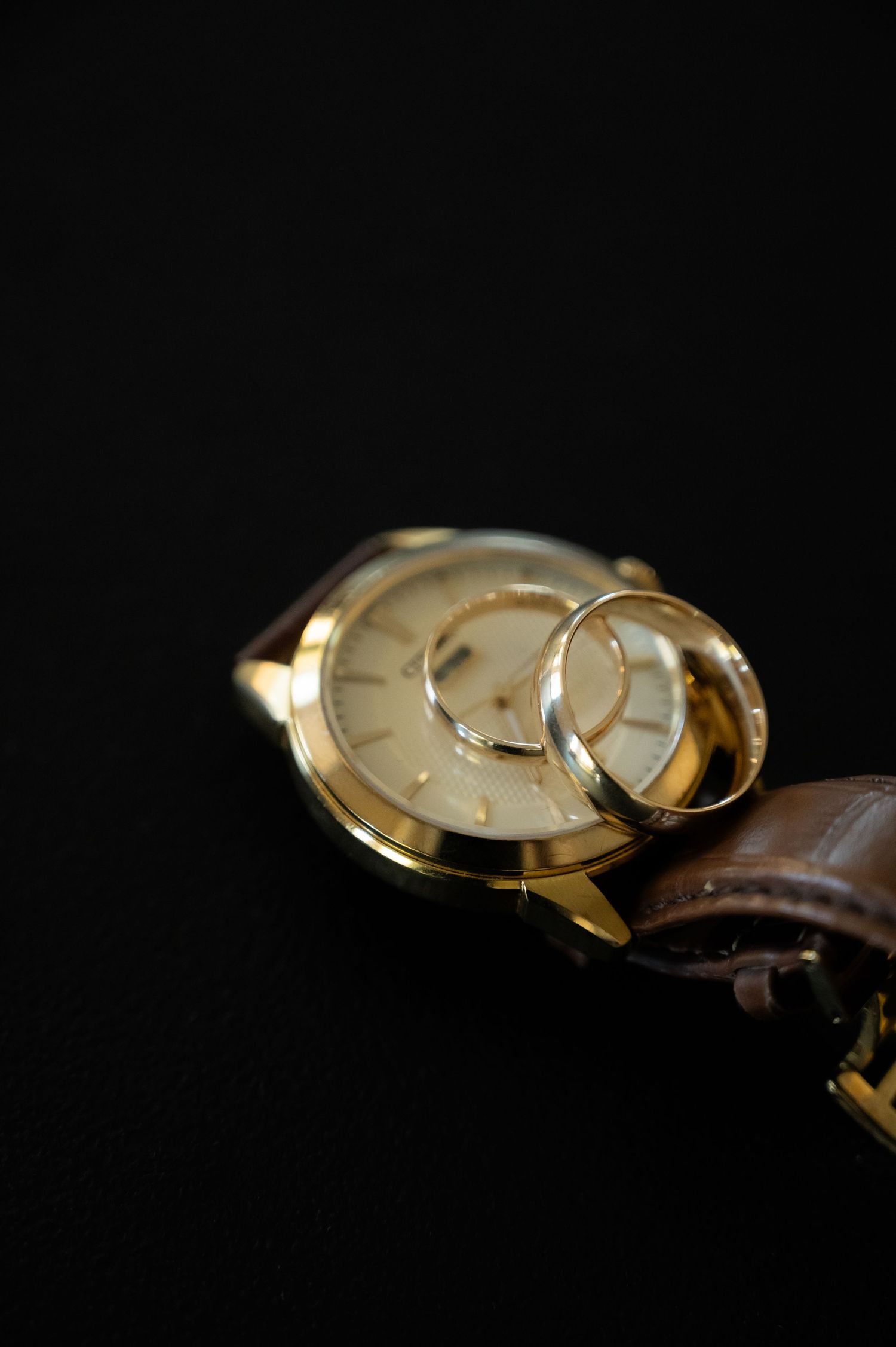 Gold watch and wedding rings arranged artistically on dark background.