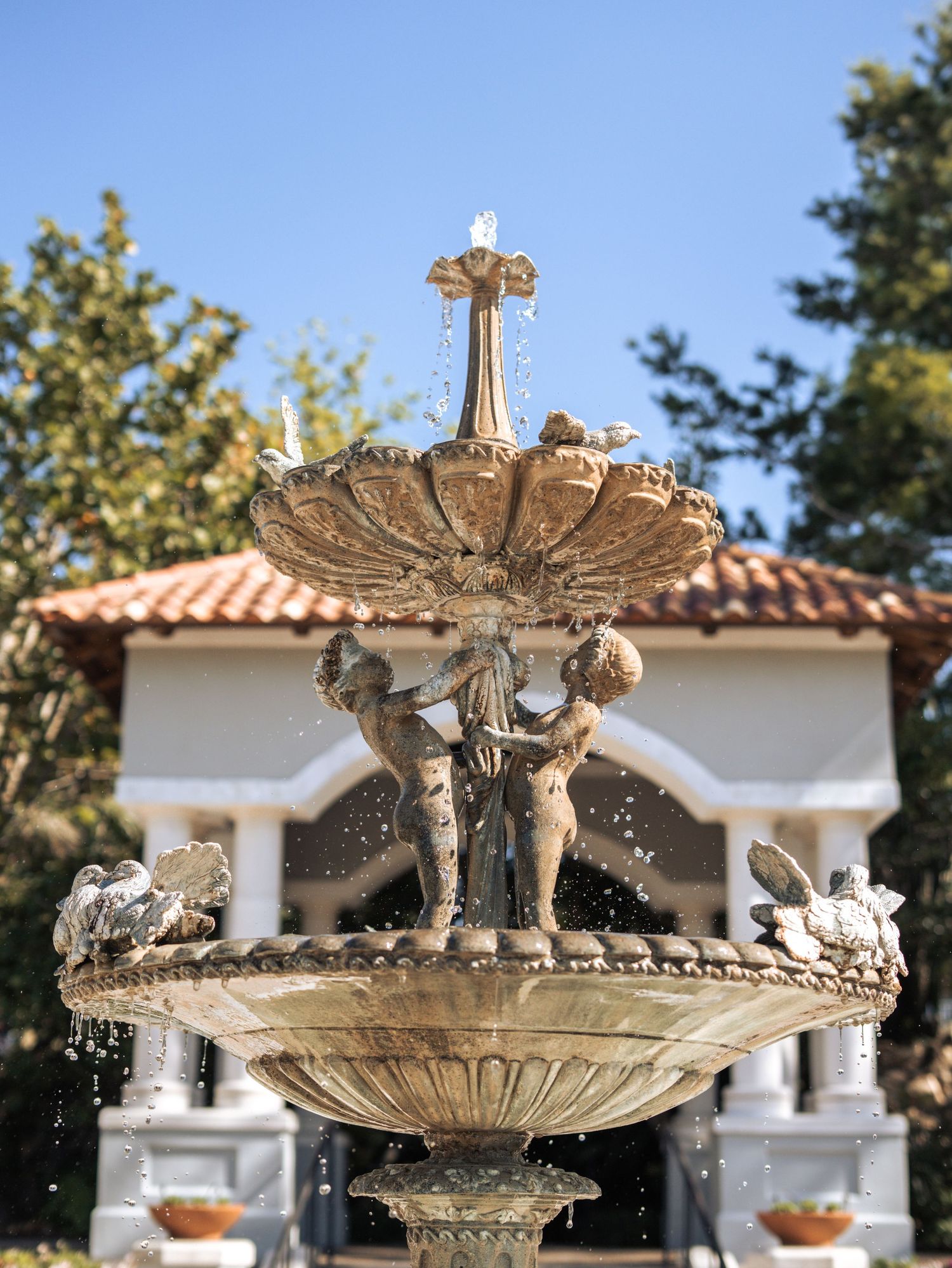 Ornate stone fountain with cherub figures spraying water in front of a Mediterranean-style building with terracotta roof tiles.