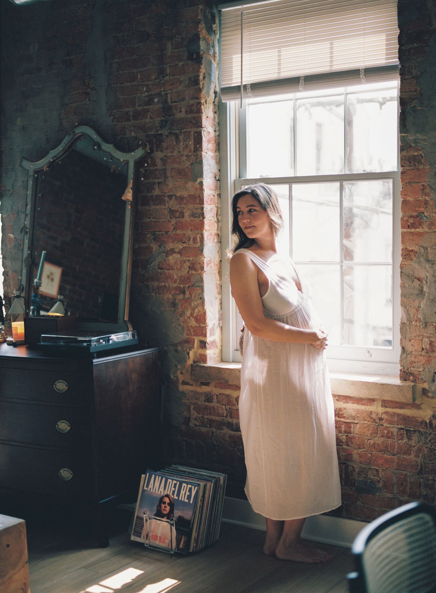 A light-filled bedroom with exposed brick walls, floral bedding, and a clothing rack visible in the corner by a turquoise window.