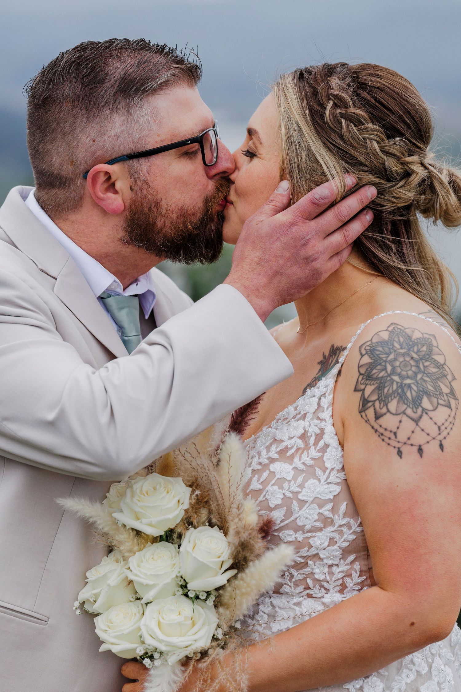 A romantic wedding kiss featuring white roses and a lace dress with a visible shoulder mandala tattoo.