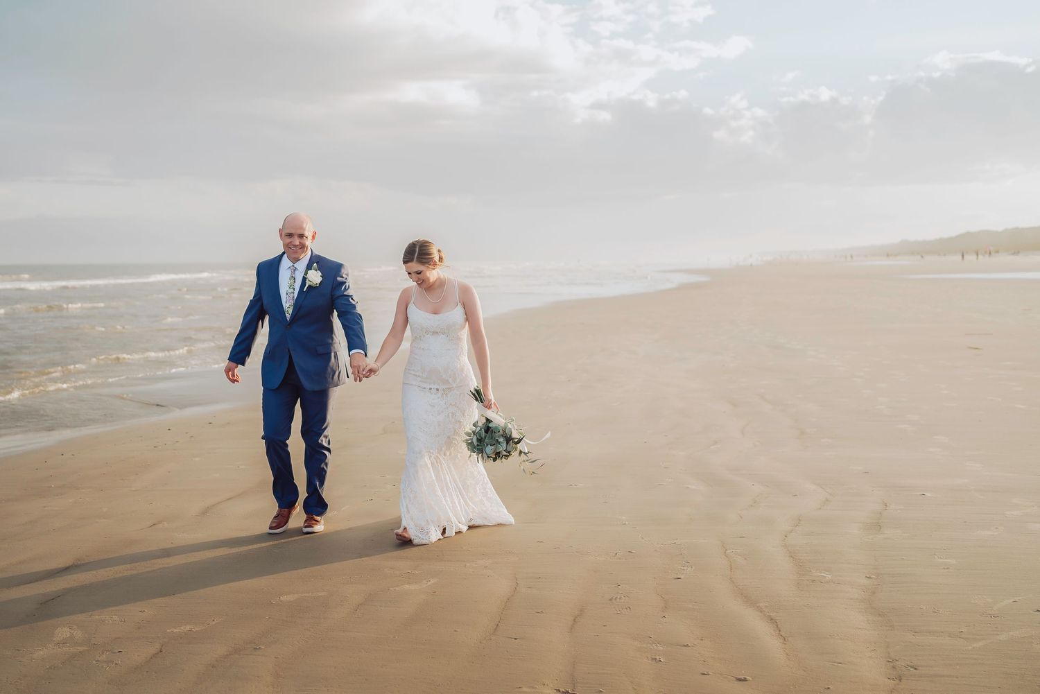 A couple in wedding attire walks hand in hand along a misty beach with waves crashing on the sand.