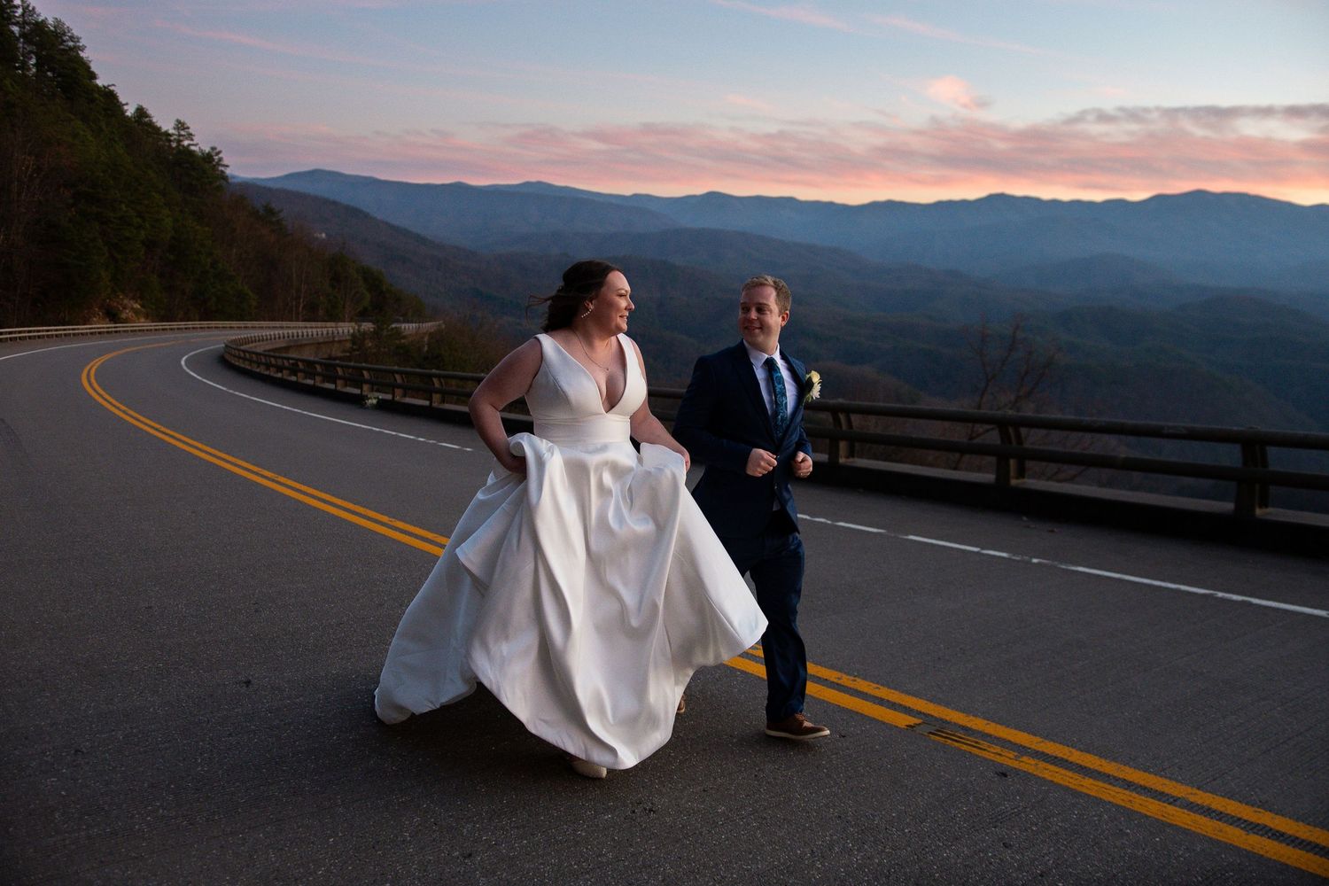 A couple shares a romantic moment on a scenic mountain road at sunset with rolling hills in the background.