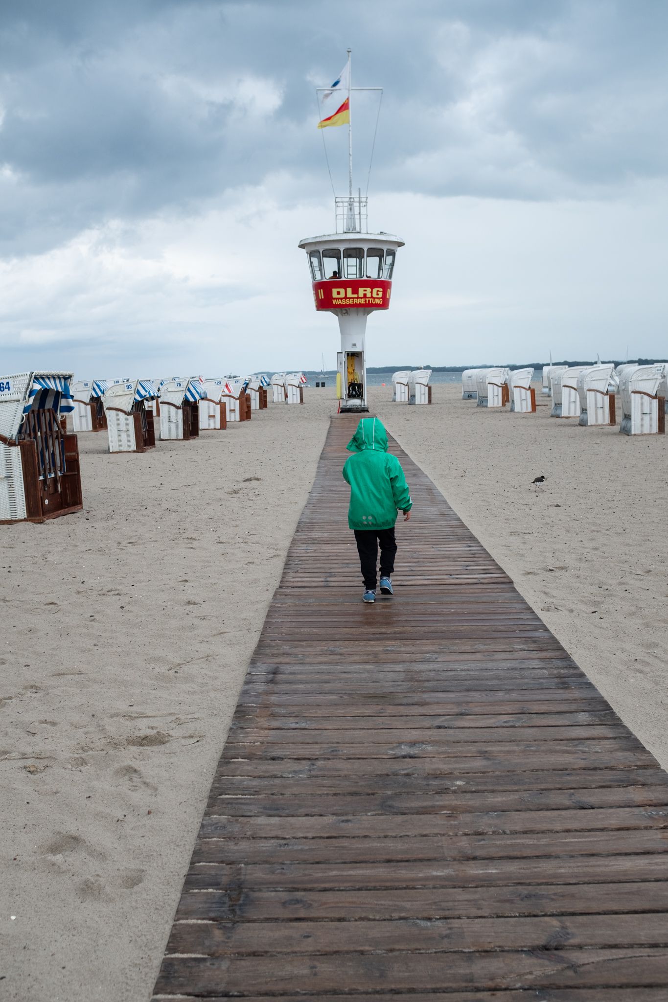 A person in green walks along a wooden boardwalk towards a lifeguard tower on a cloudy beach with colorful changing huts.
