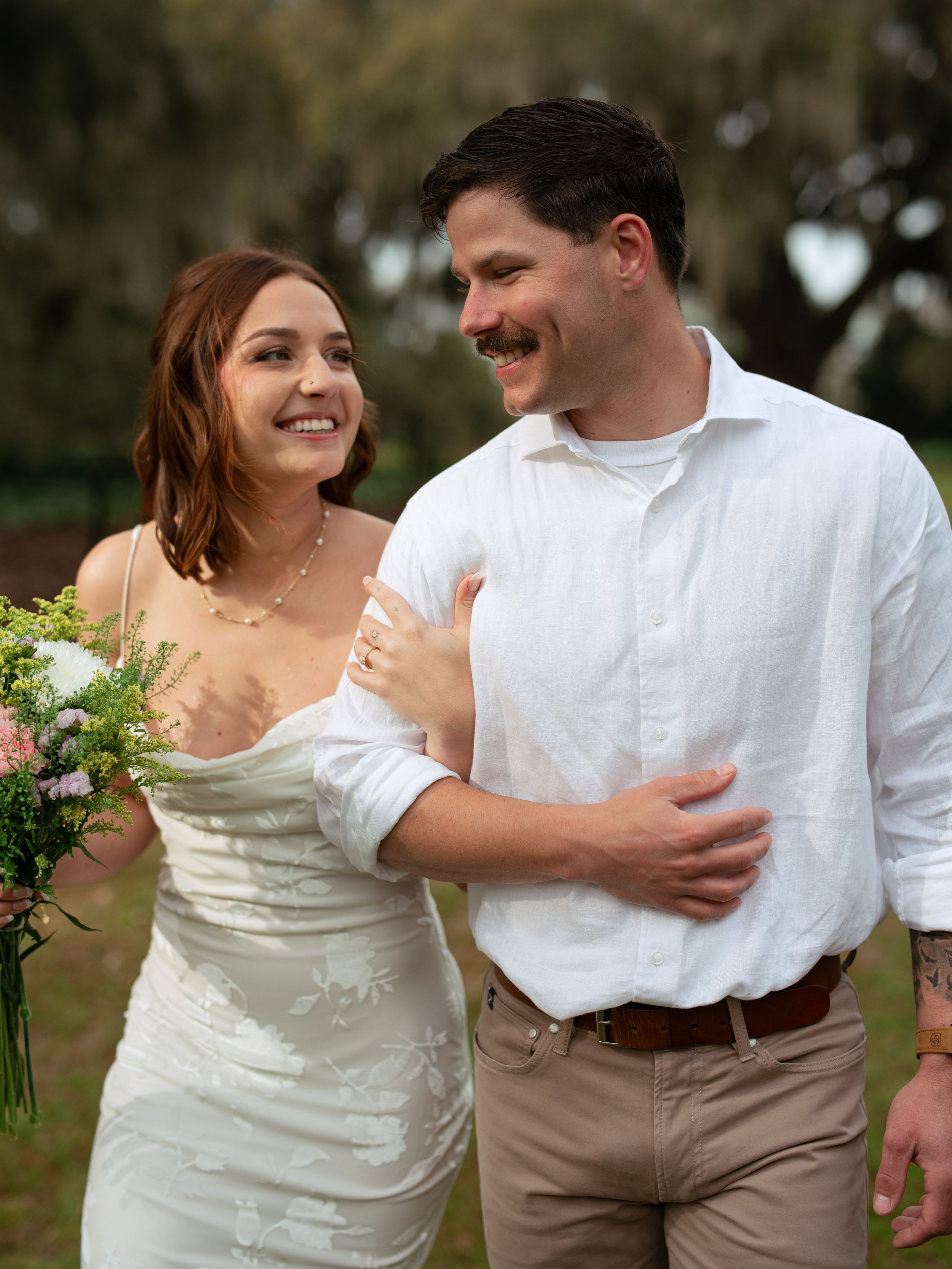 A couple shares a happy moment outdoors at their wedding, with the bride wearing a white dress and holding a bouquet.