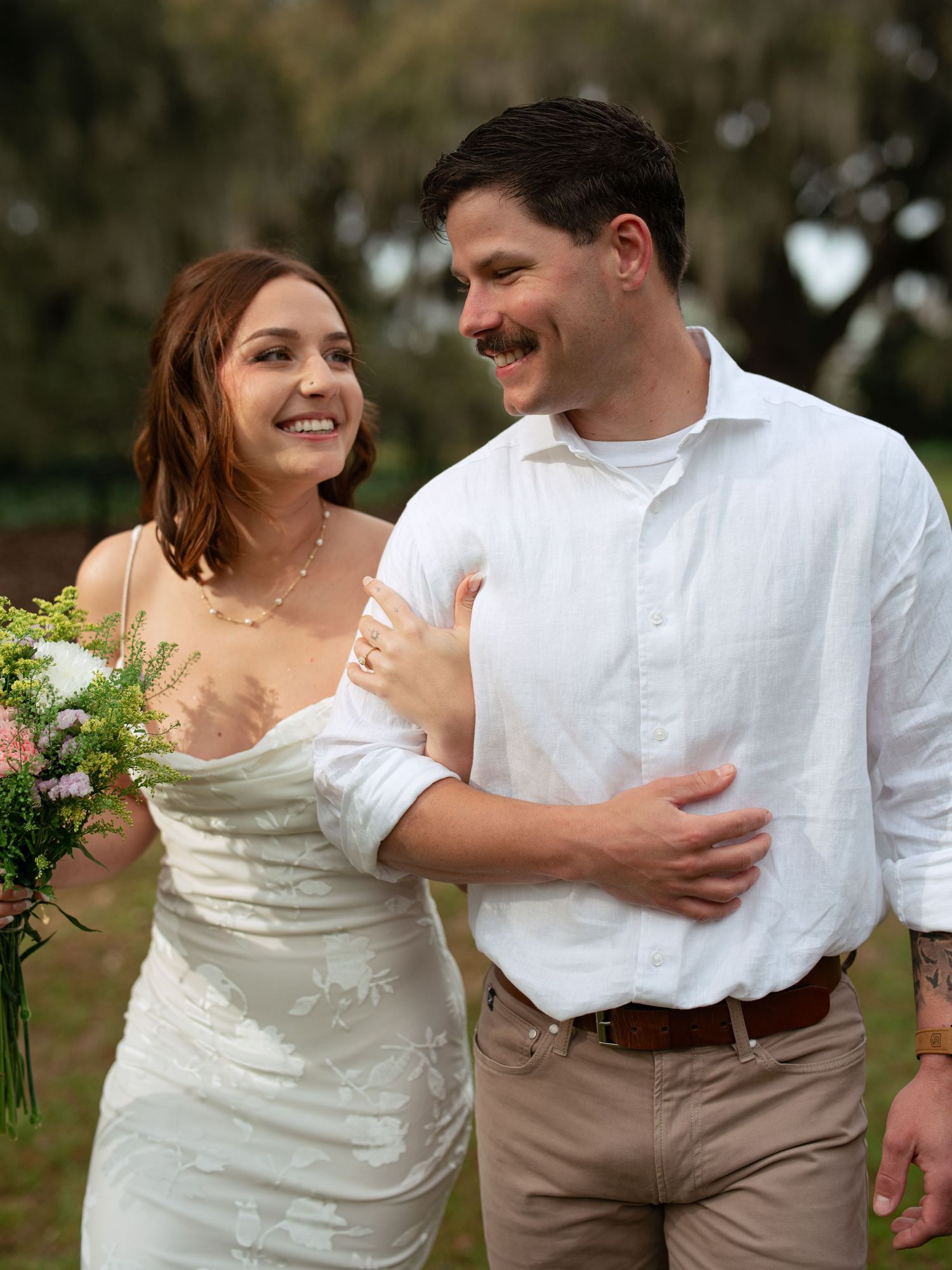 A couple shares a happy moment outdoors at their wedding, with the bride wearing a white dress and holding a bouquet.