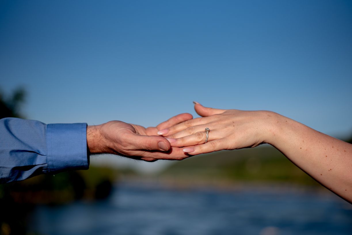 Two hands reach towards each other against a blue sky backdrop during a romantic engagement moment.