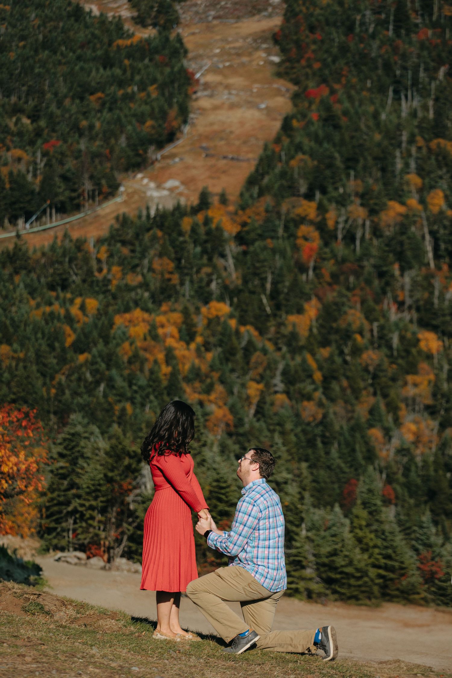 A couple shares a romantic proposal moment on a mountainside trail surrounded by autumn foliage and evergreen trees.