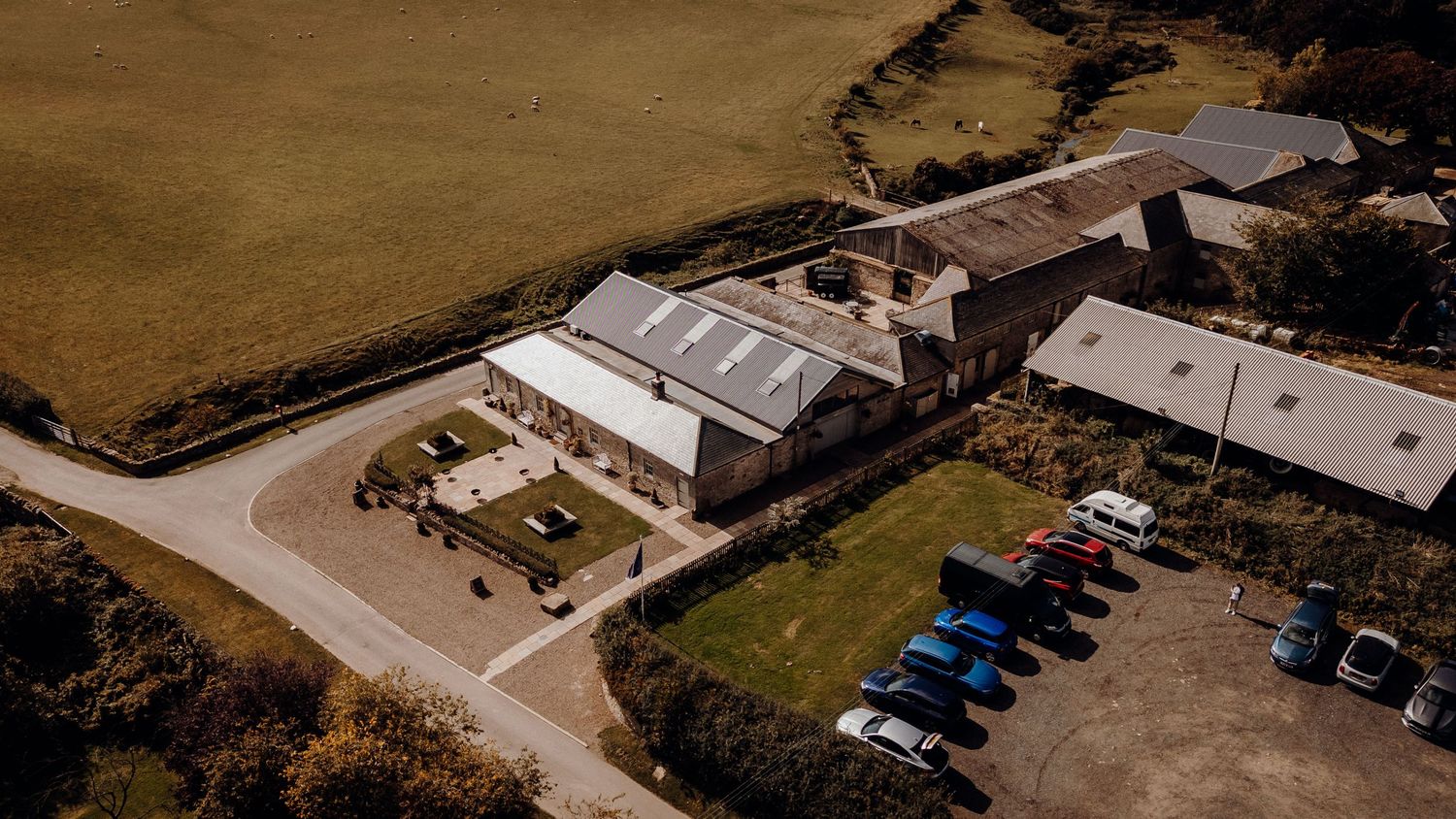 Aerial view of industrial buildings with white roofs and a parking area containing blue vehicles in a rural setting.