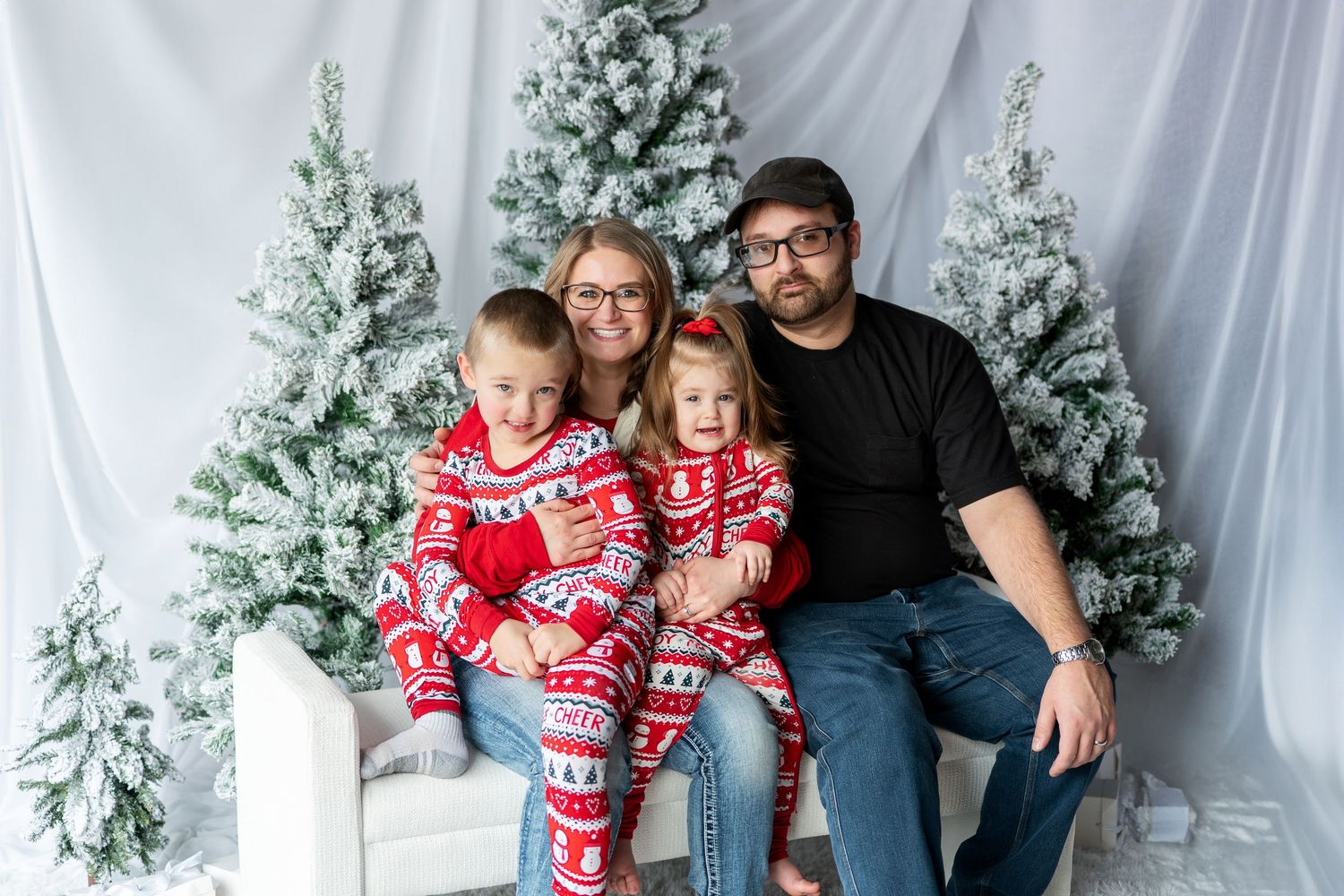 Family of four in matching red holiday pajamas posing together with snow-flocked Christmas trees in a studio.