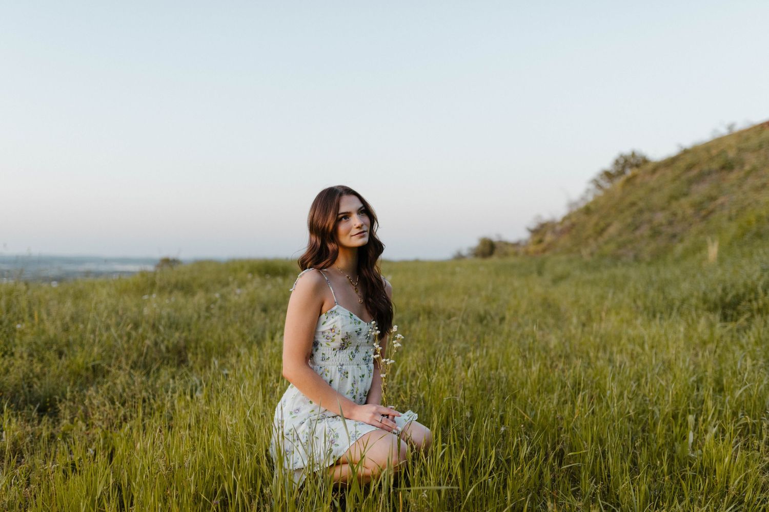 A figure in a floral dress poses in a sunlit meadow with coastal views in the background.