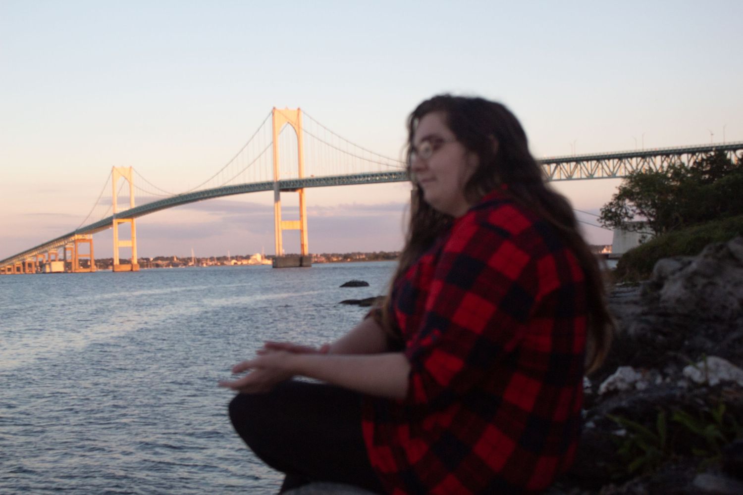 Someone in a red plaid flannel shirt sits by the Newport Bridge at sunset over Narragansett Bay in Rhode Island.