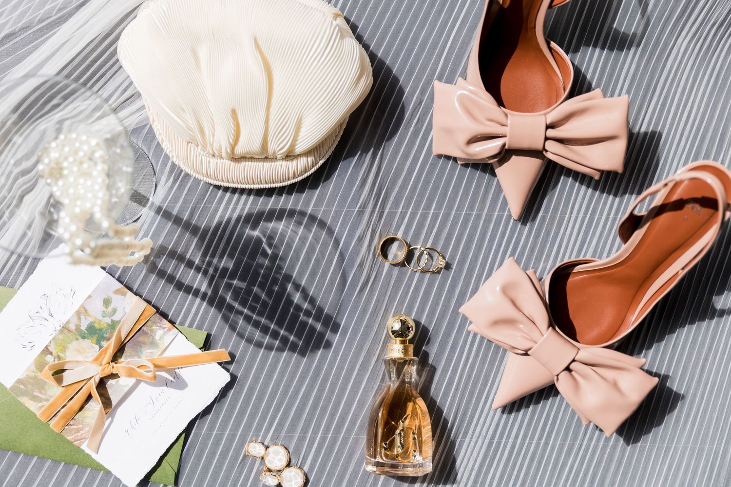 Flat lay styling with pink bow flats, perfume bottle, and wedding accessories on gray fabric background.