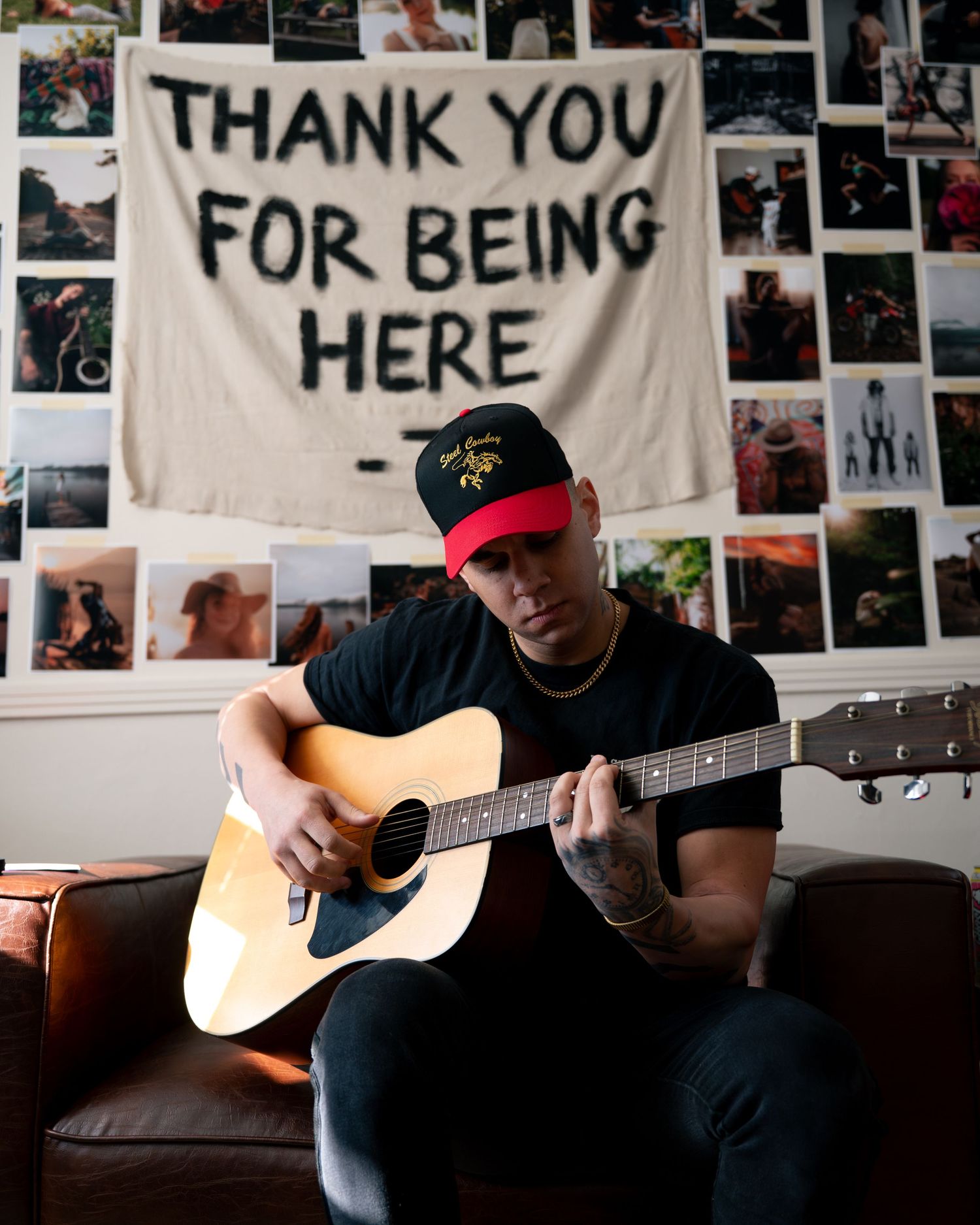 Musician in dark clothing plays acoustic guitar in room decorated with photographs and banners on the wall.