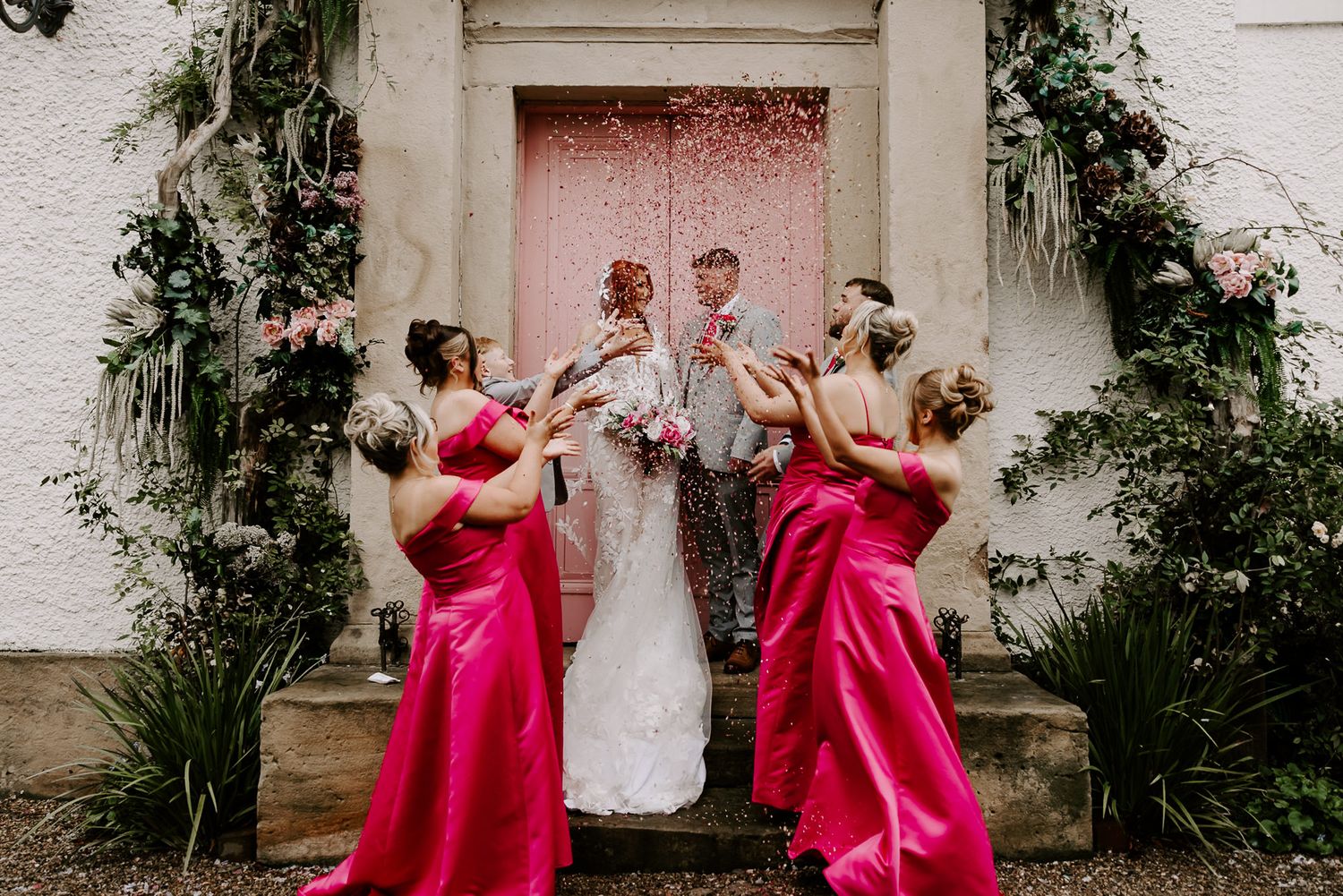 A bridal party in bright pink dresses poses playfully during a wedding photo session at a white building exterior.