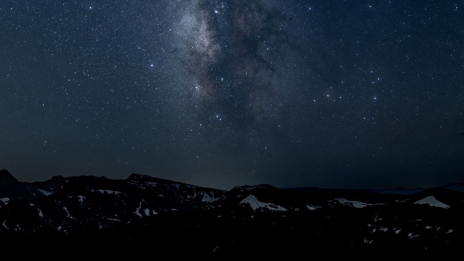The Milky Way galaxy stretches across a dark night sky above snow-capped mountain peaks in a stunning starscape above Rocky Mt National Park