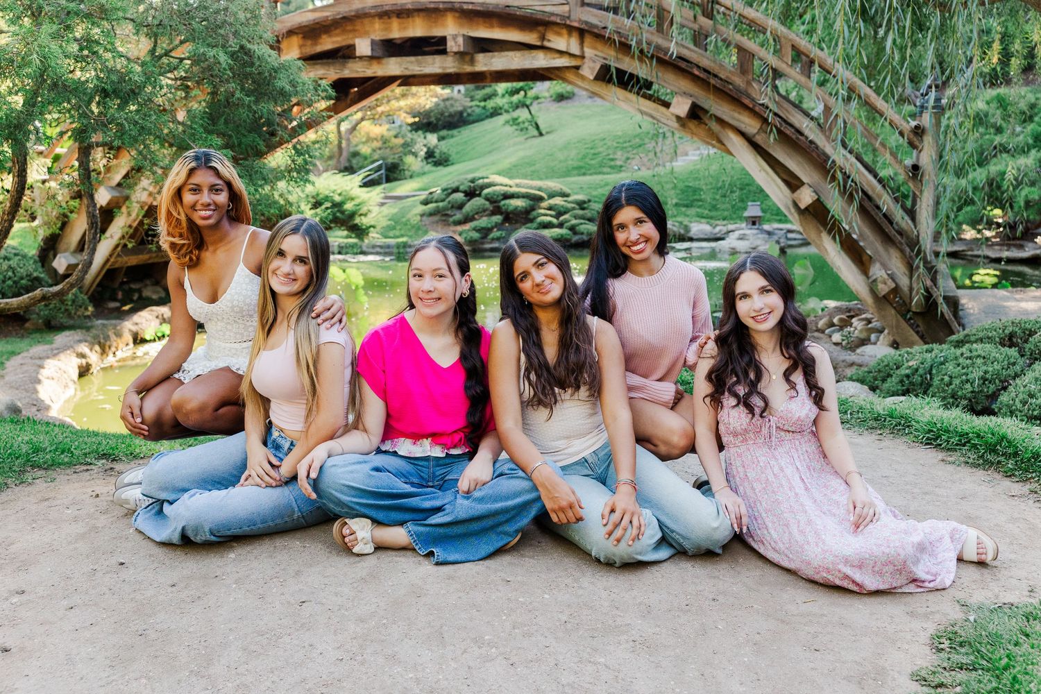 A group of people sit together and laugh on a wooden bridge in a lush garden setting.