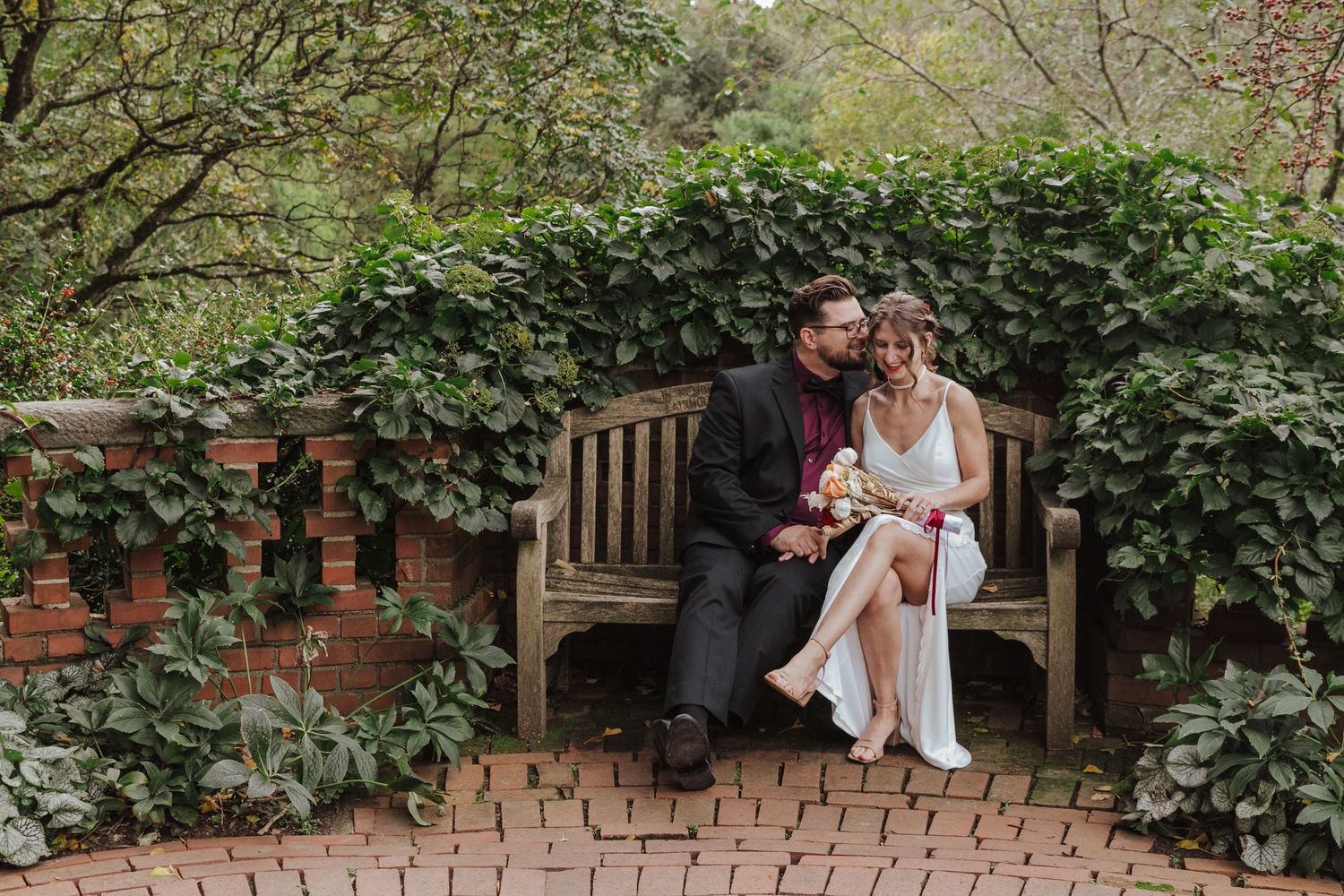 A couple sits together on a wooden garden bench surrounded by lush greenery and brick pathways in a garden setting.