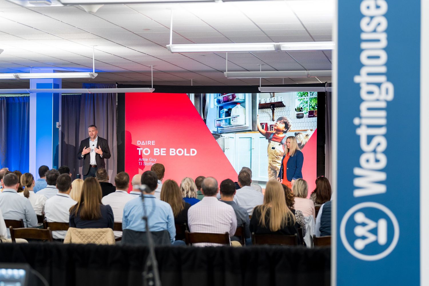 A speaker presents on stage at a business conference with blue curtain backdrop and audience in silhouette.