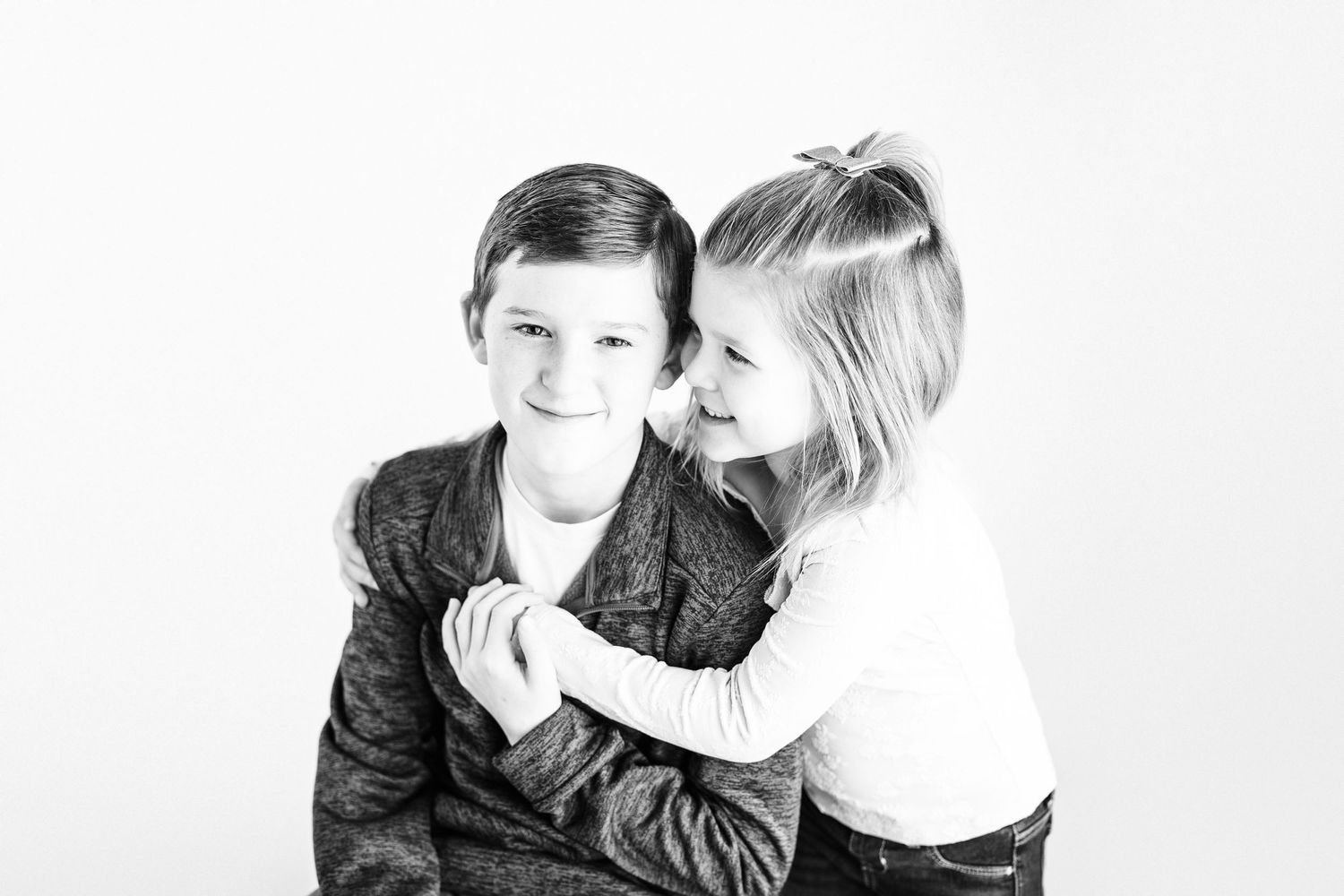 Black and white studio portrait of two children sharing a playful moment against a white background.