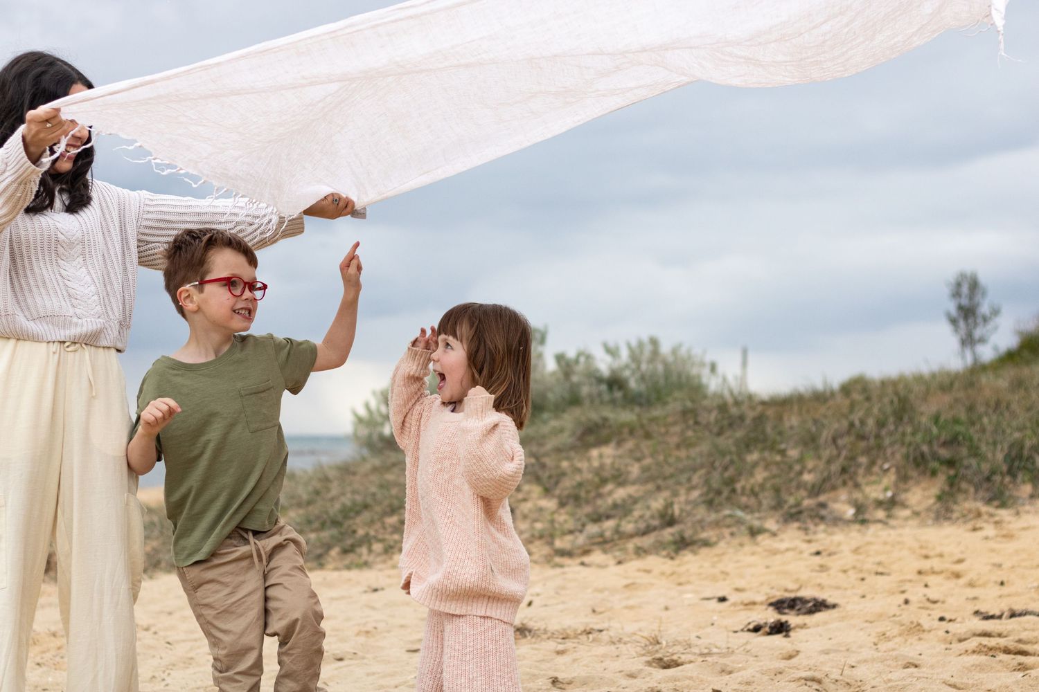 A family plays together on a sandy beach with white fabric blowing in the wind on a cloudy day.