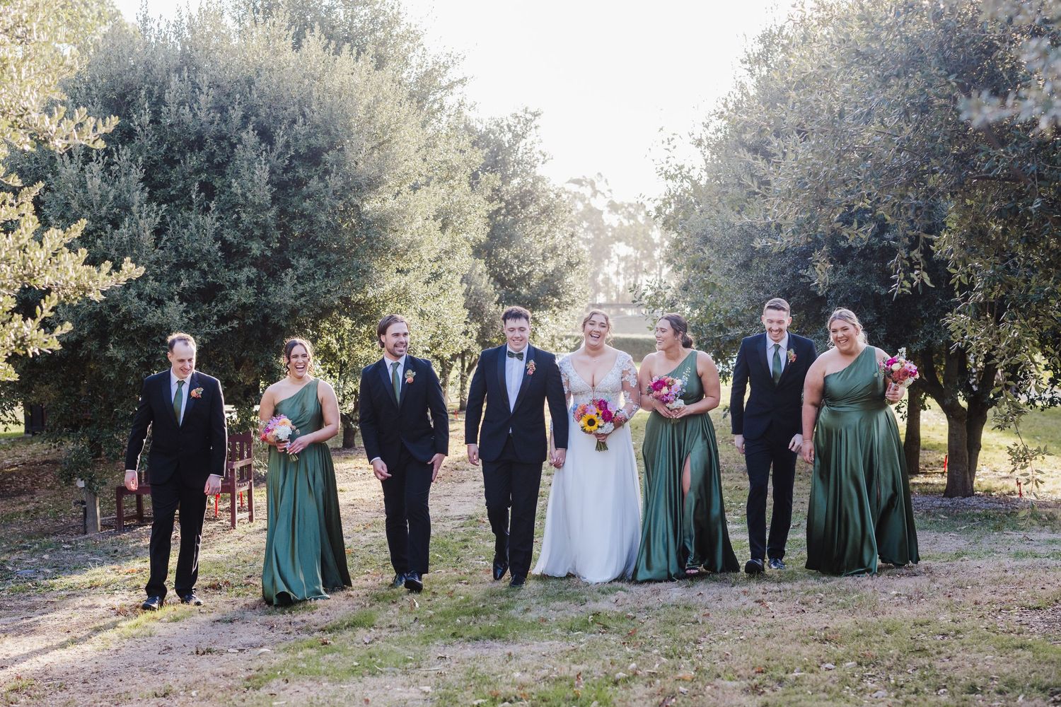 A couple walks together along a sunlit path between rows of trees at an outdoor wedding ceremony.