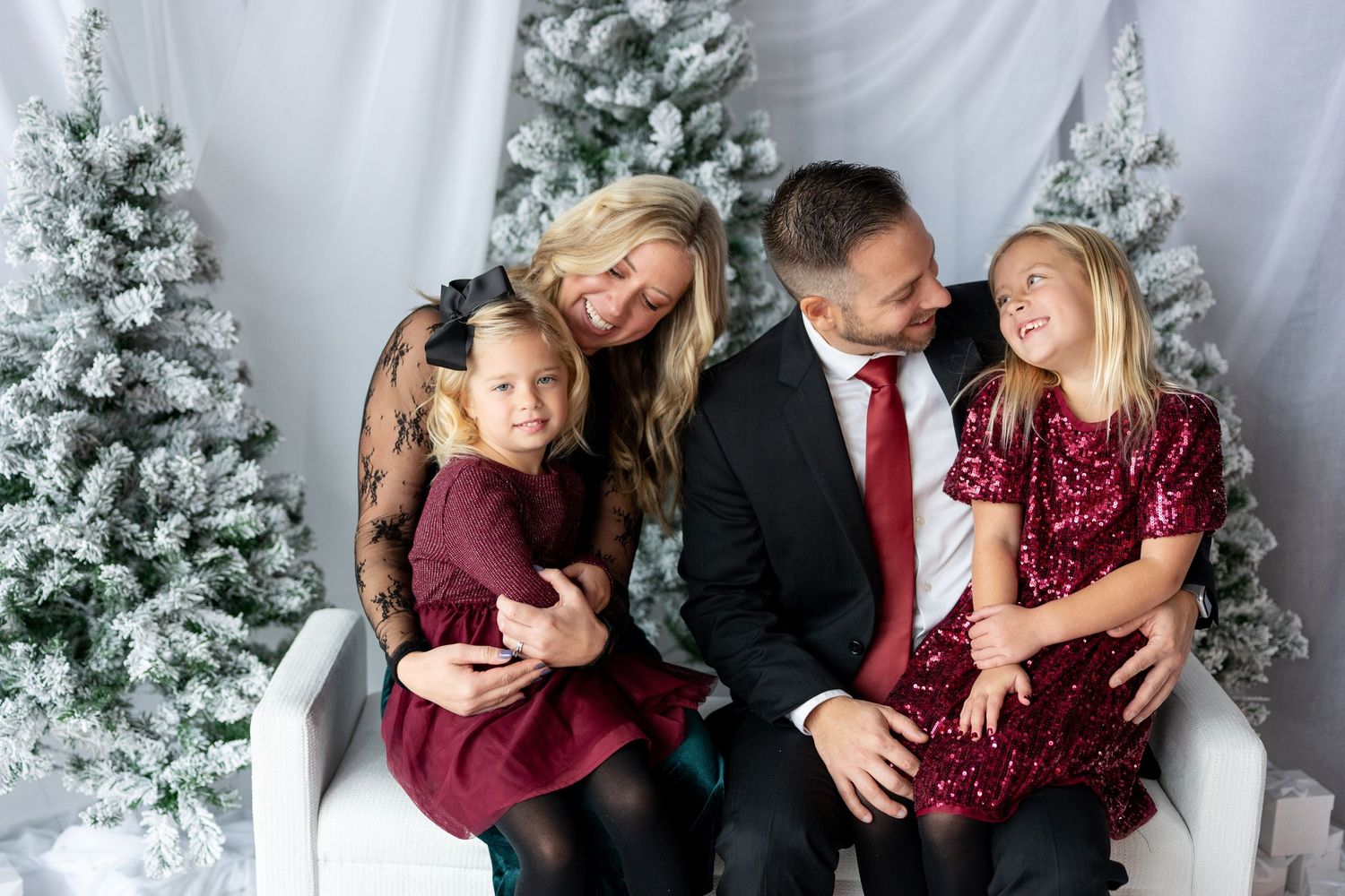 Happy family of four posing together on a white bench surrounded by snow-flocked Christmas trees in festive attire.