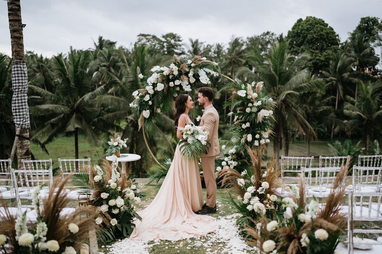 Outdoor wedding venue decorated with white flowers and chairs among palm trees.