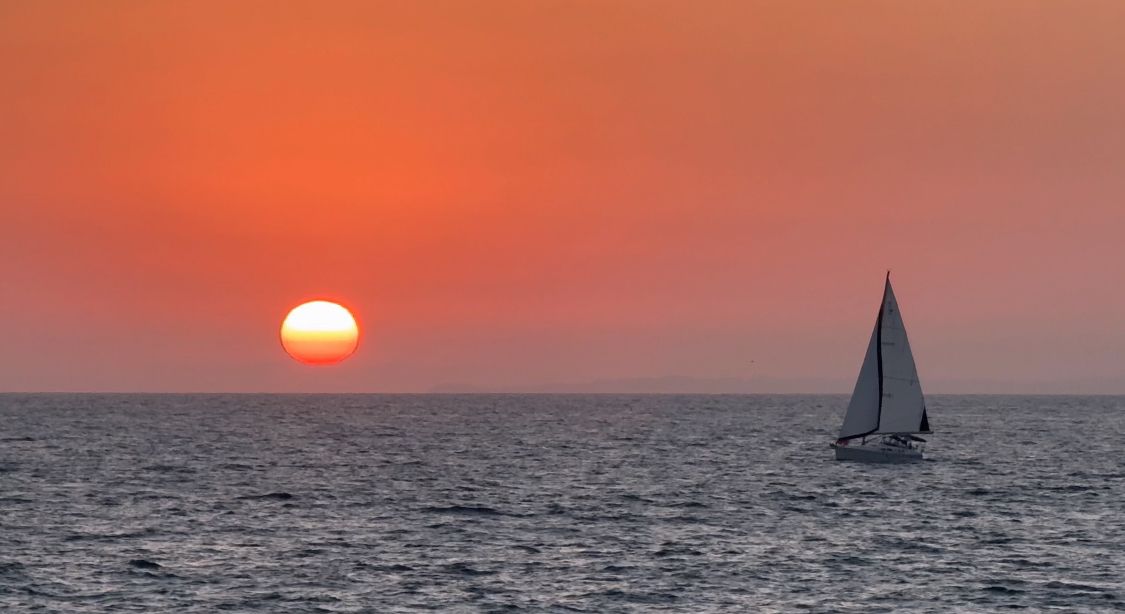 A sailboat silhouetted against a vibrant orange sunset sky over calm ocean waters.