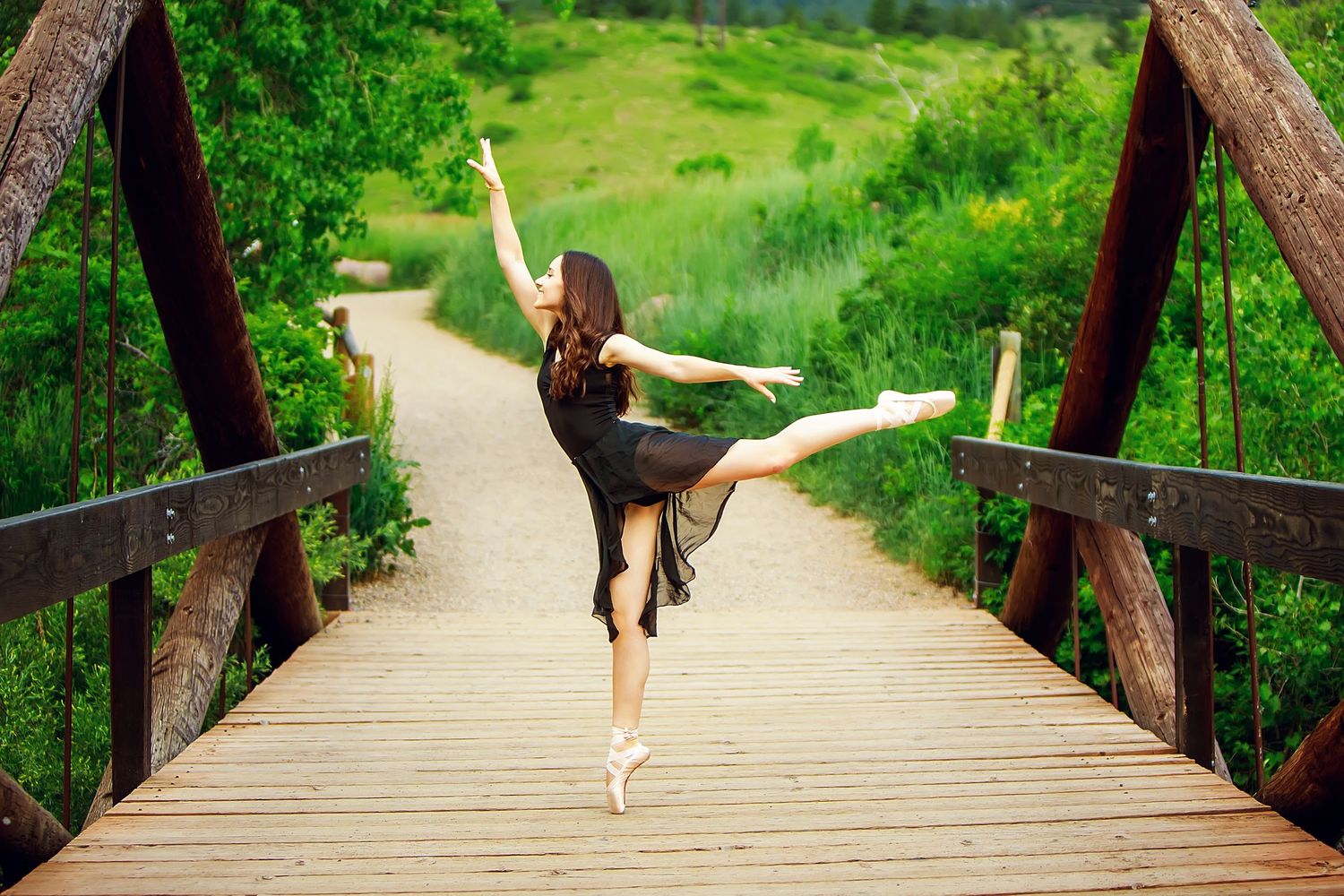 A graceful ballet dancer performs an arabesque pose on a wooden bridge surrounded by lush green scenery.