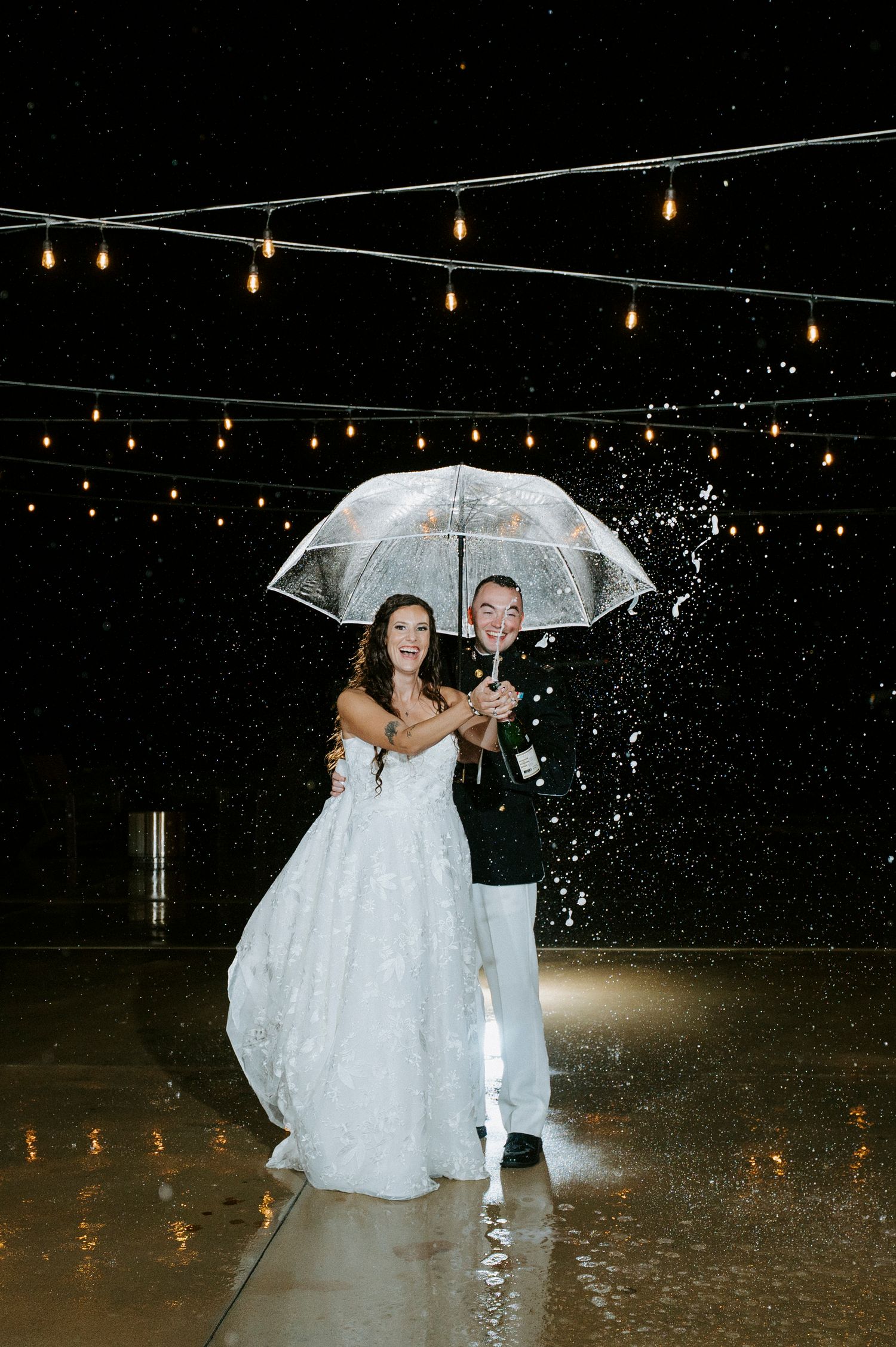 A couple shares a romantic moment under a clear umbrella in the rain with string lights glowing overhead.