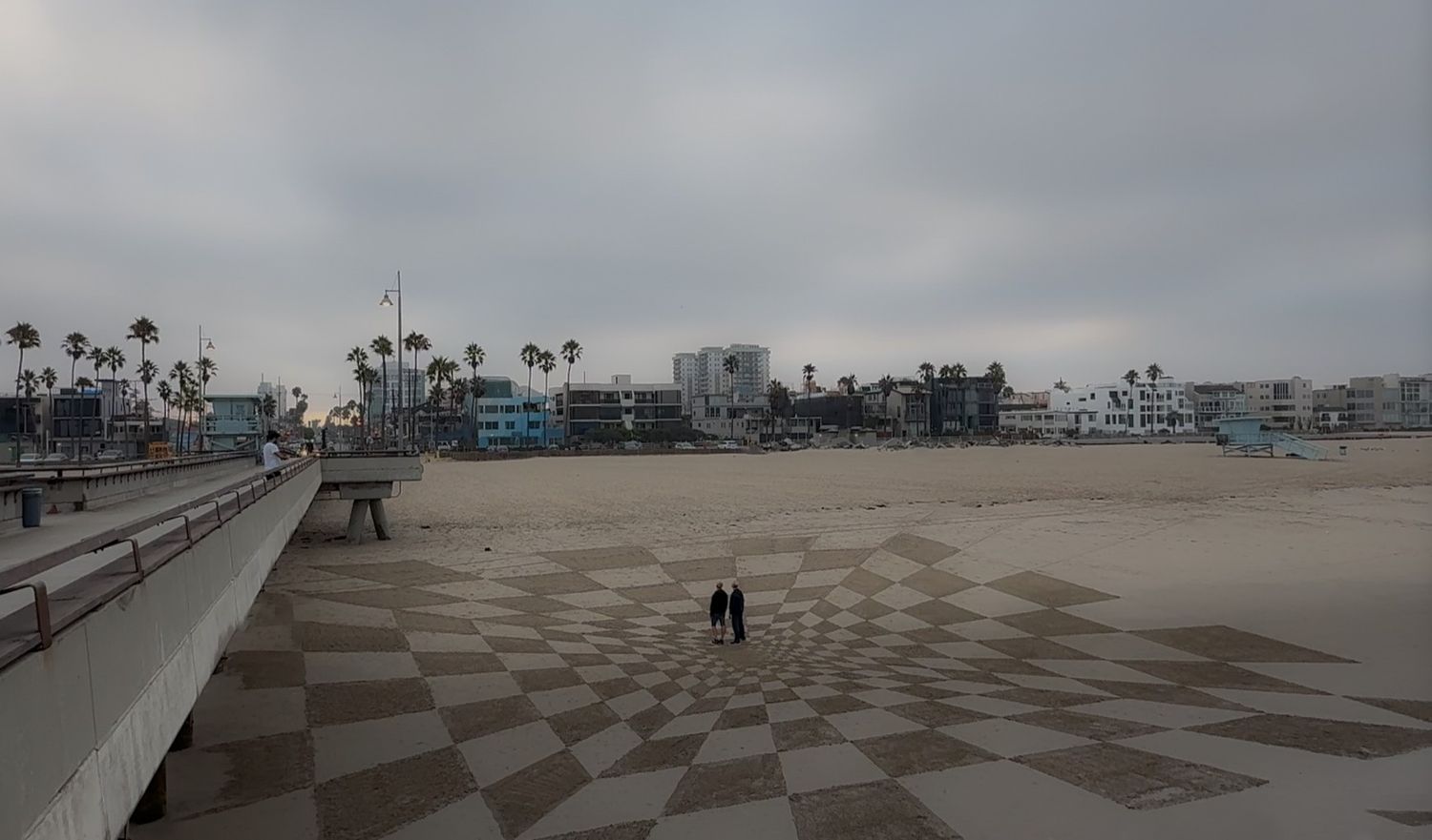 Moody beach scene with checkered cement walkways and palm trees along the Venice Beach boardwalk on a cloudy day.