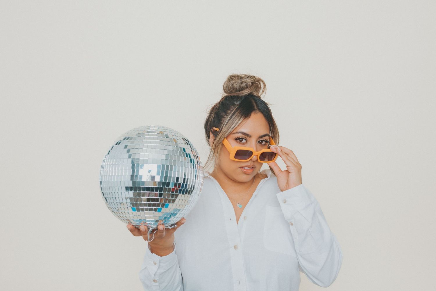 A person in a white sweater holds a silver disco ball while posing against a light colored background.
