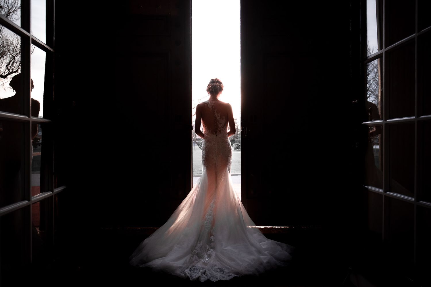 Silhouette of a bride in a flowing mermaid wedding gown standing in a doorway with dramatic backlighting.