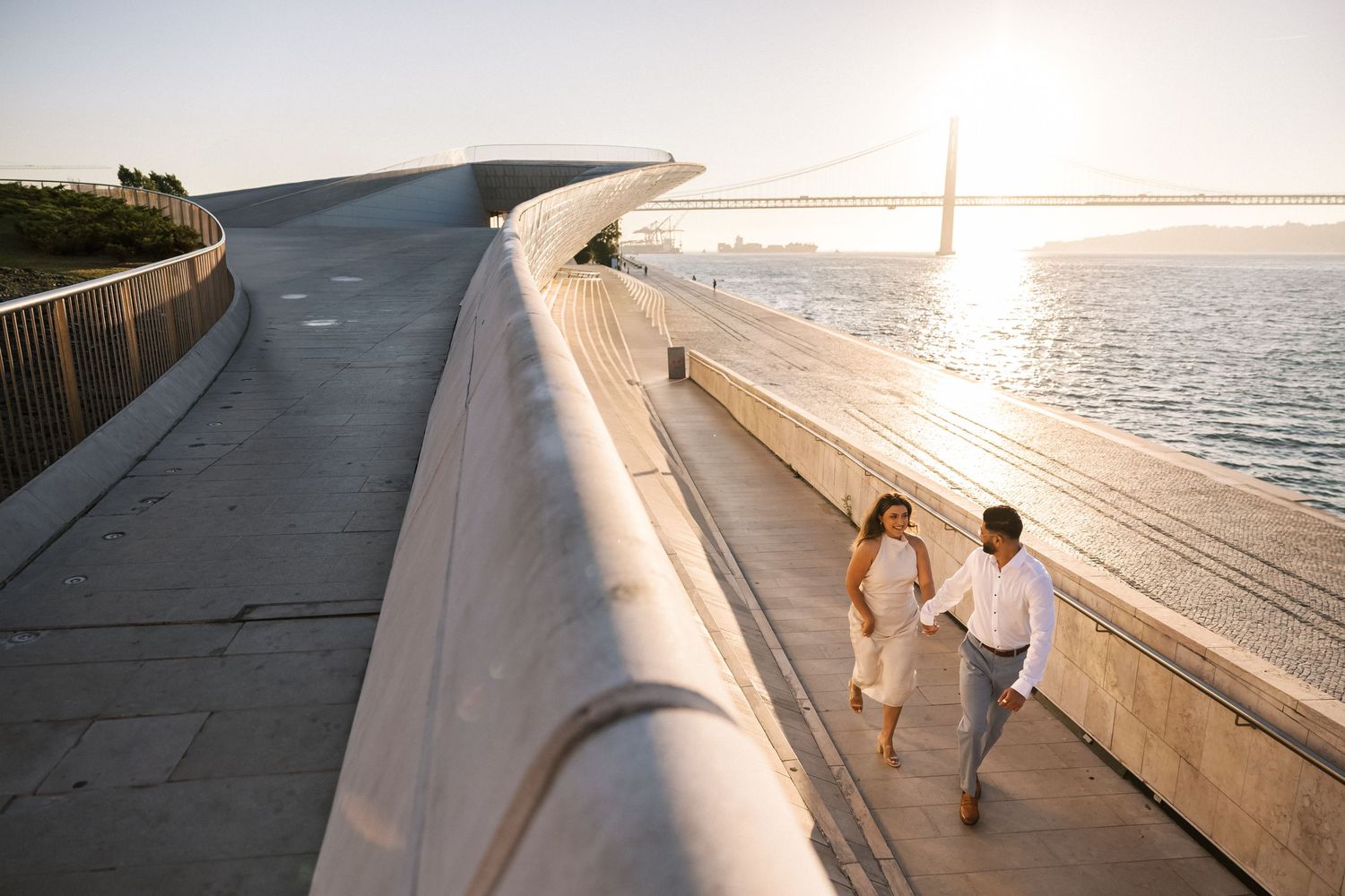Couple walking hand in hand along Lisbon riverfront at sunrise, with 25 de Abril Bridge in the background — engagement session in Portugal.