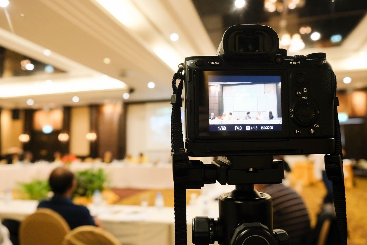 Digital camera on tripod recording an event in a conference room with blurred background and bokeh lighting.
